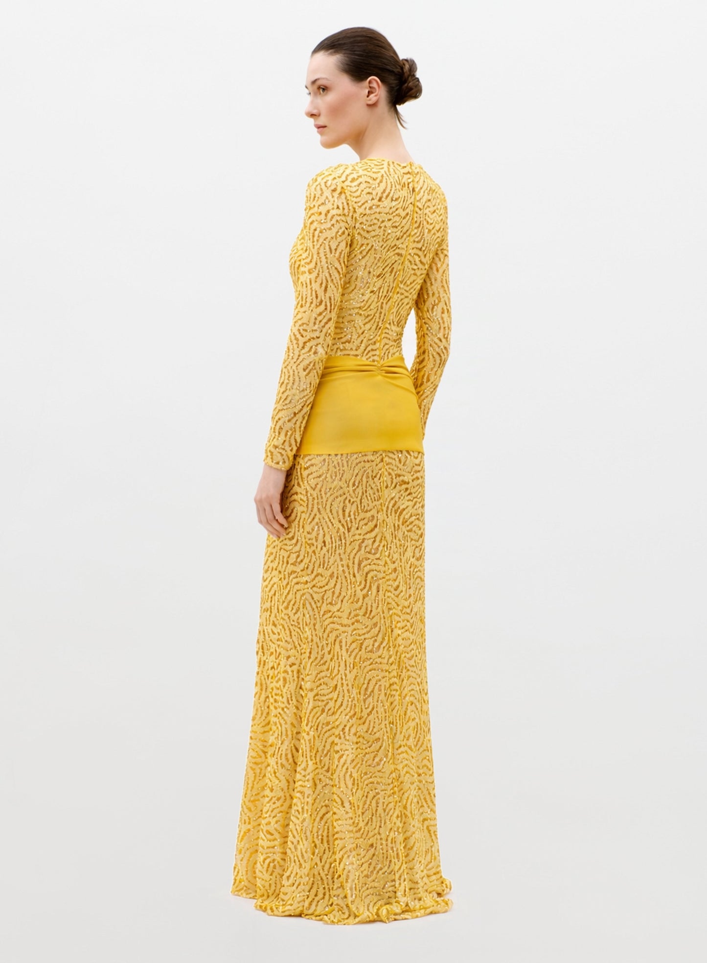 A woman with her hair in a neat bun wears the Lia Dress—a long-sleeved, floor-length yellow dress featuring a textured pattern and solid yellow waistband—standing against a plain light background.