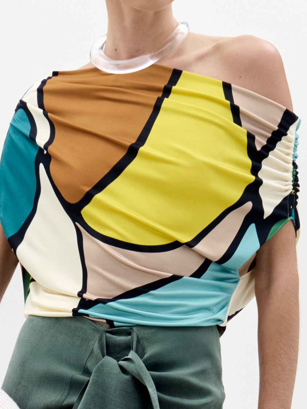 A woman stands against a plain background wearing the colorful Indiana Blouse with an off-the-shoulder neckline, a green high-waisted skirt with a bow, and holding a white textured bag.
