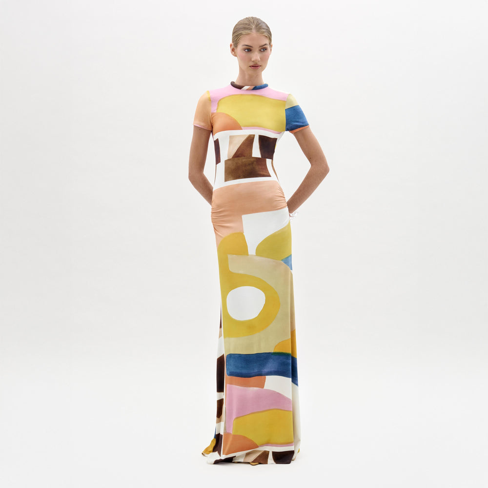 A woman stands against a white background wearing the Ikia Dress, a floor-length, short-sleeved maxi crafted from Italian fabric in a colorful abstract pattern of yellow, pink, blue, brown, and white shapes.