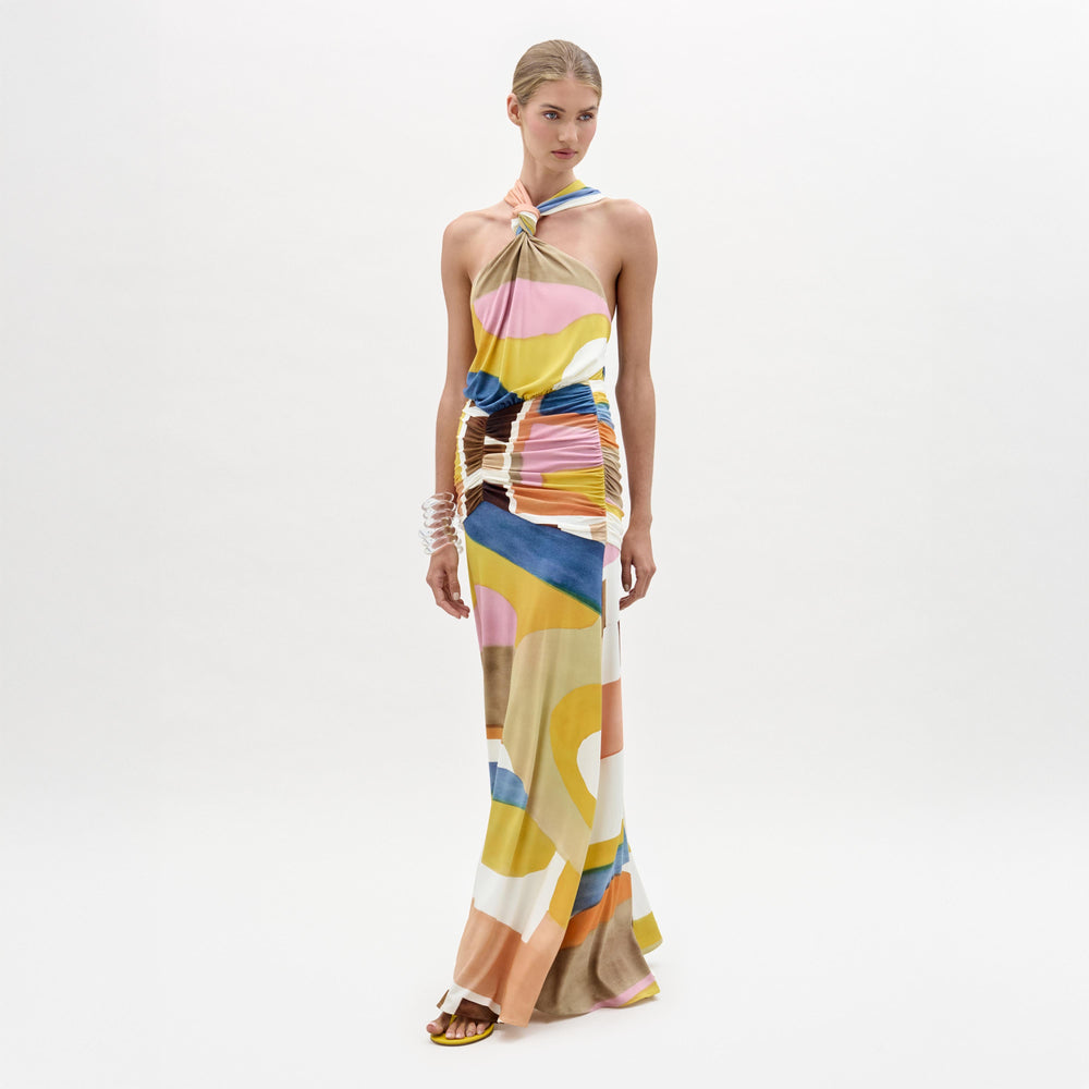 A woman stands against a white background wearing the Jedda Dress, a colorful abstract-patterned maxi with a halter neck, ruched details, and strappy sandals.
