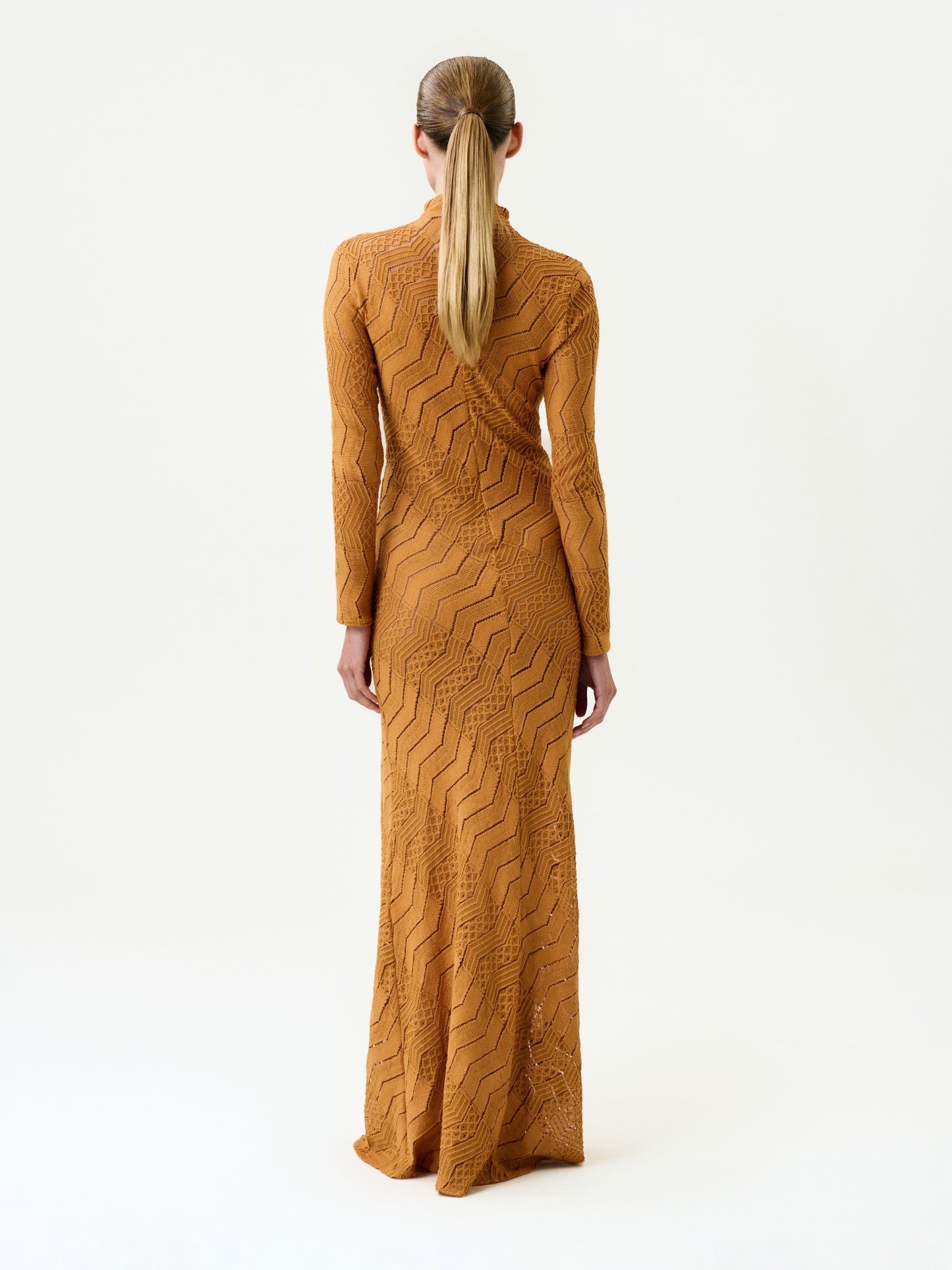A person with long, straight hair in a low ponytail wears the Marianne Dress—a floor-length amber maxi featuring a zigzag texture, made in Colombia—standing against a plain white background, facing away from the camera.