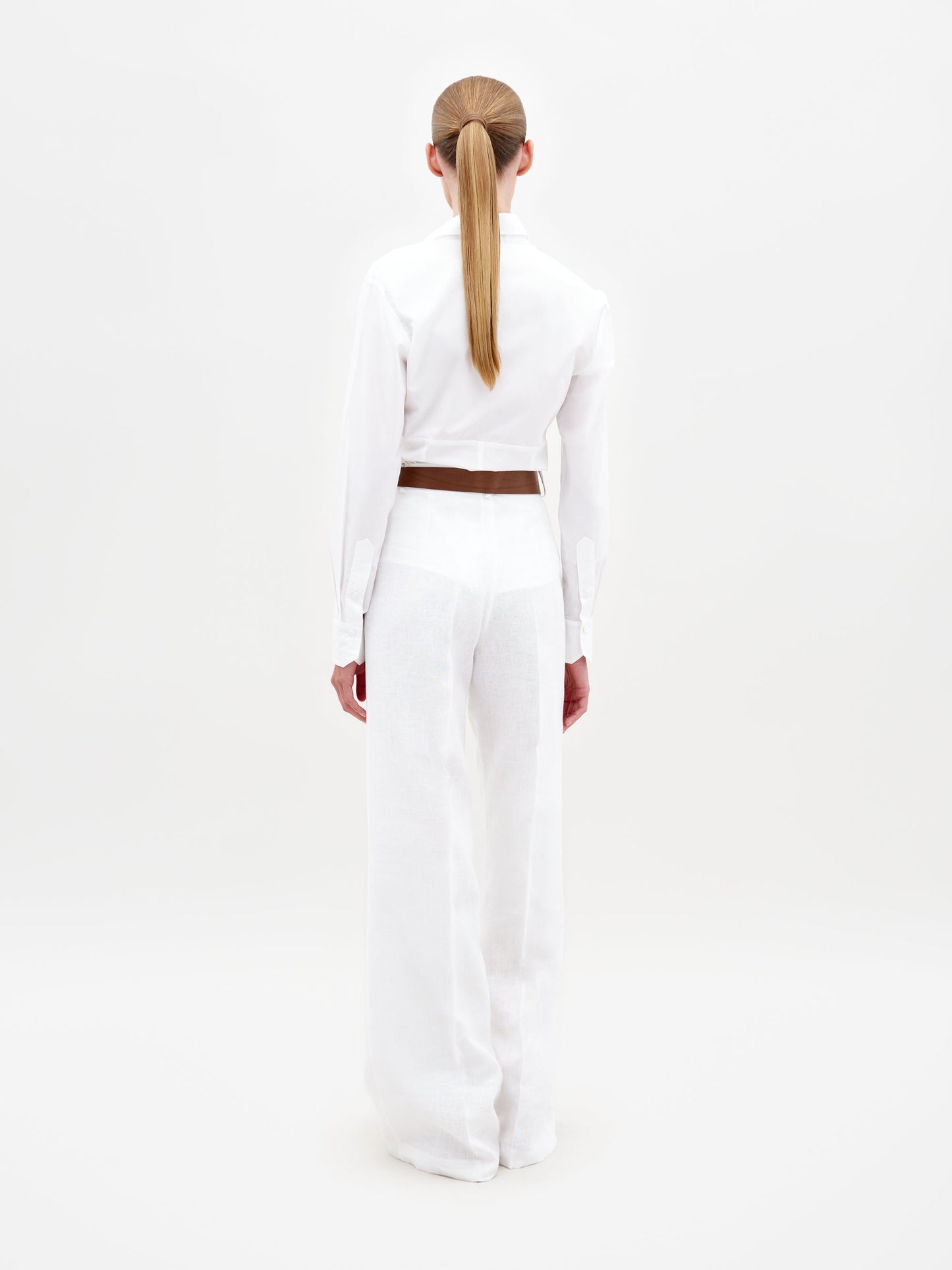 A person with long, straight hair in a low ponytail stands with their back to the camera, wearing the Sigrid Blouse, wide-leg white pants, and a brown belt against a plain white background.