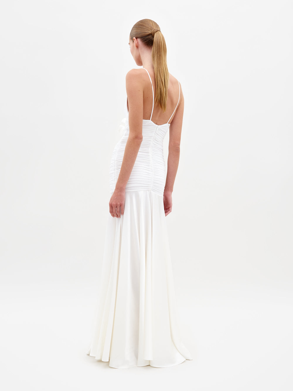 A woman in the Sana Dress White, featuring a sleeveless design with a deep V-neck and floral detail at the waist, stands against a plain white background. Pre-order now to ensure delivery by February 15th, 2025.