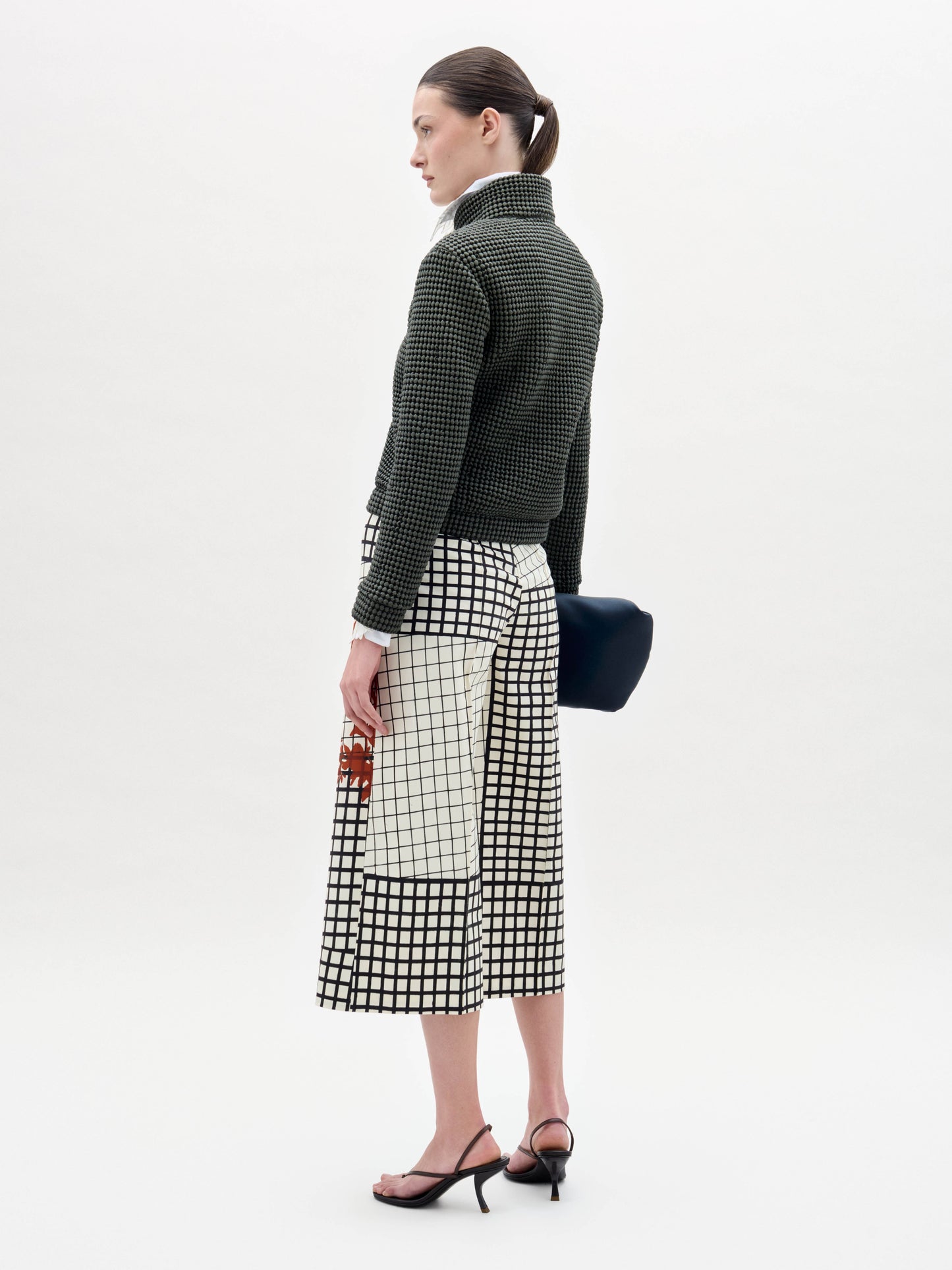 A woman with a neutral expression stands against a plain background wearing the textured Zalta Jacket, patterned pants, and holding a dark bag.