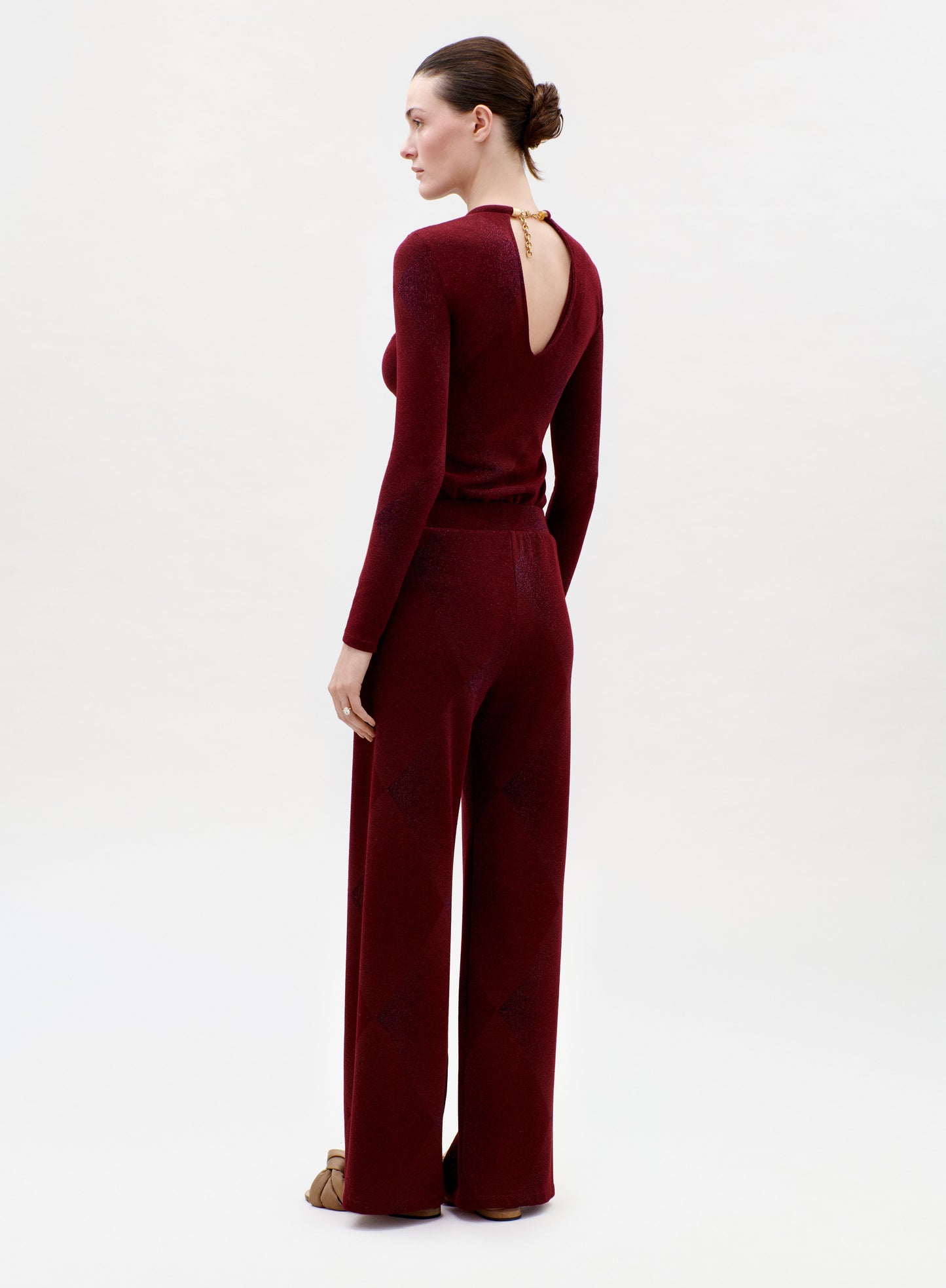 A woman stands facing away in the Colbie Pant and a maroon long-sleeve top with a back keyhole cutout. Her hair is in a neat bun, she wears brown shoes, and the background is plain and light.