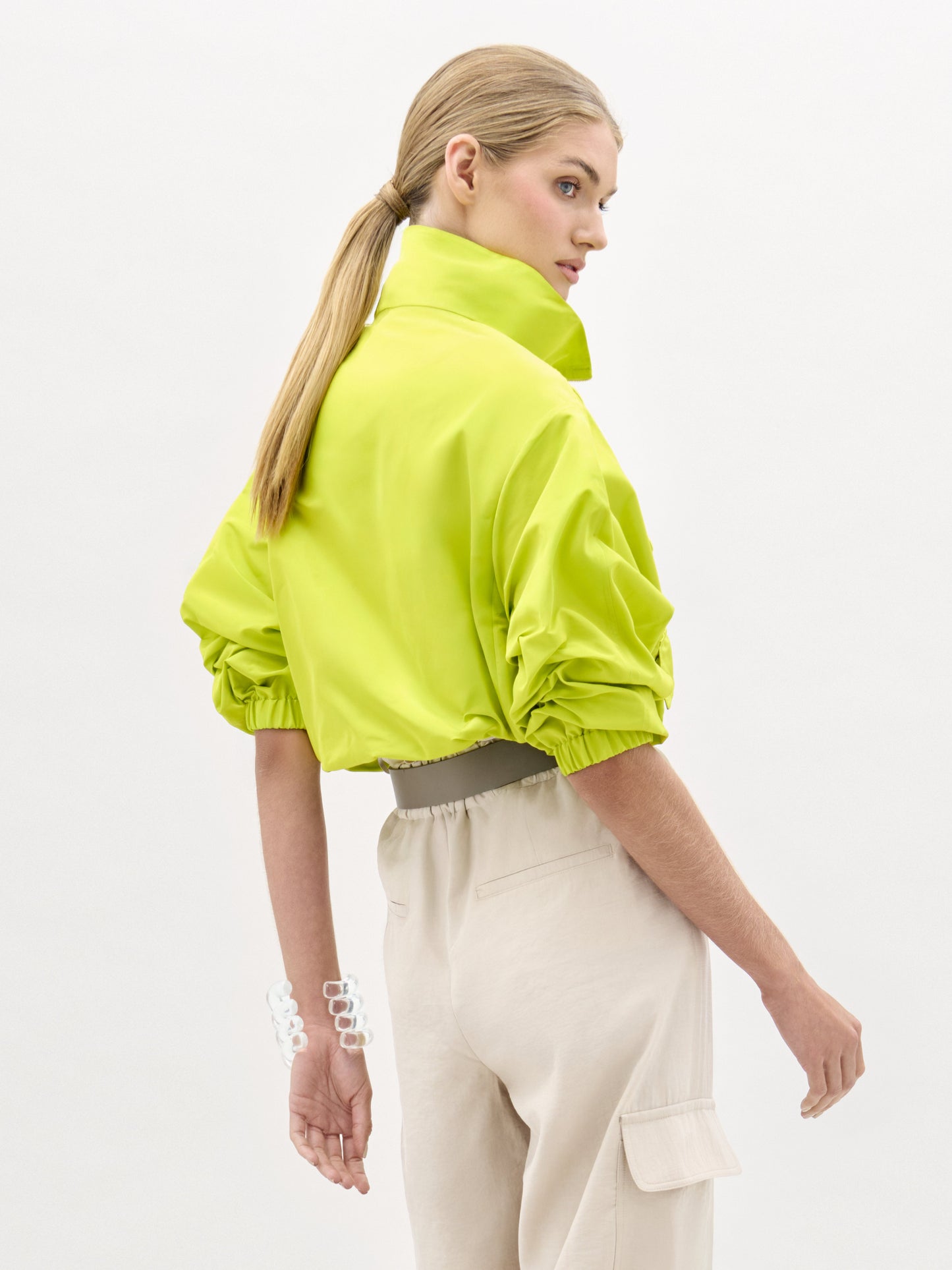 A woman wears the Donata Jacket, a bright yellow cropped jacket with floral embroidery, paired with beige high-waisted pants, a wide belt featuring a circular buckle, and a clear bracelet as she stands against a plain background.