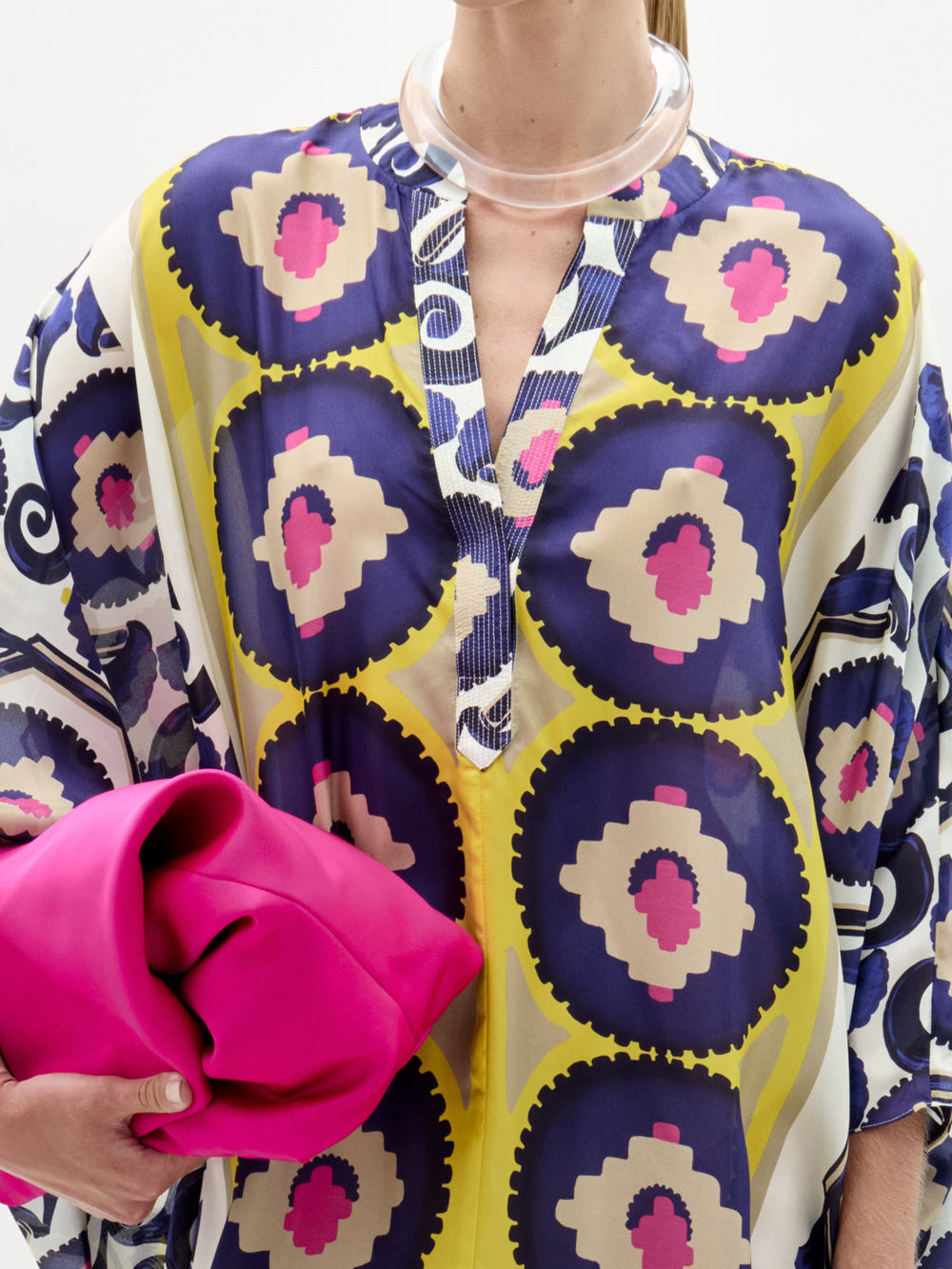 Woman models the Ilse Tunic, a long maxi dress crafted from Italian fabric featuring blue, yellow, and pink circles. She holds a bright pink clutch and wears heeled sandals against a plain white background.