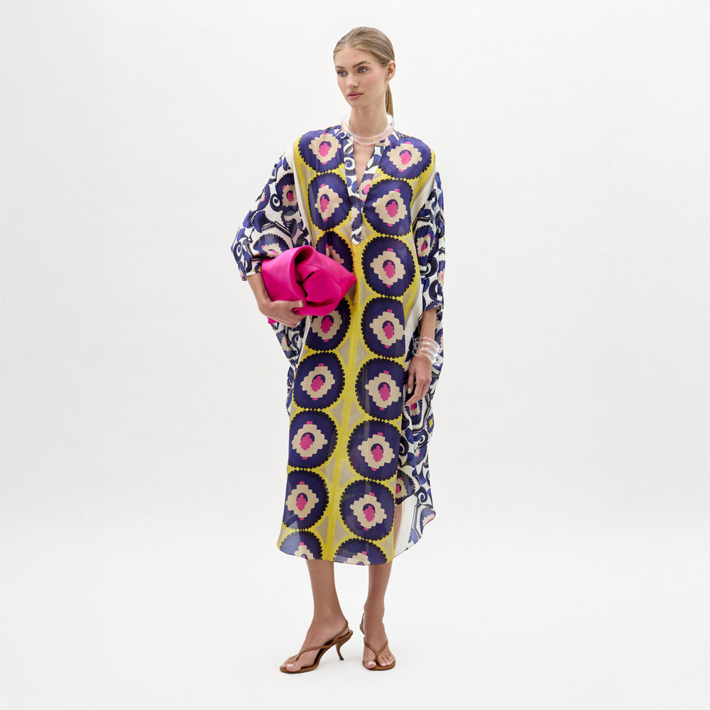 Woman models the Ilse Tunic, a long maxi dress crafted from Italian fabric featuring blue, yellow, and pink circles. She holds a bright pink clutch and wears heeled sandals against a plain white background.