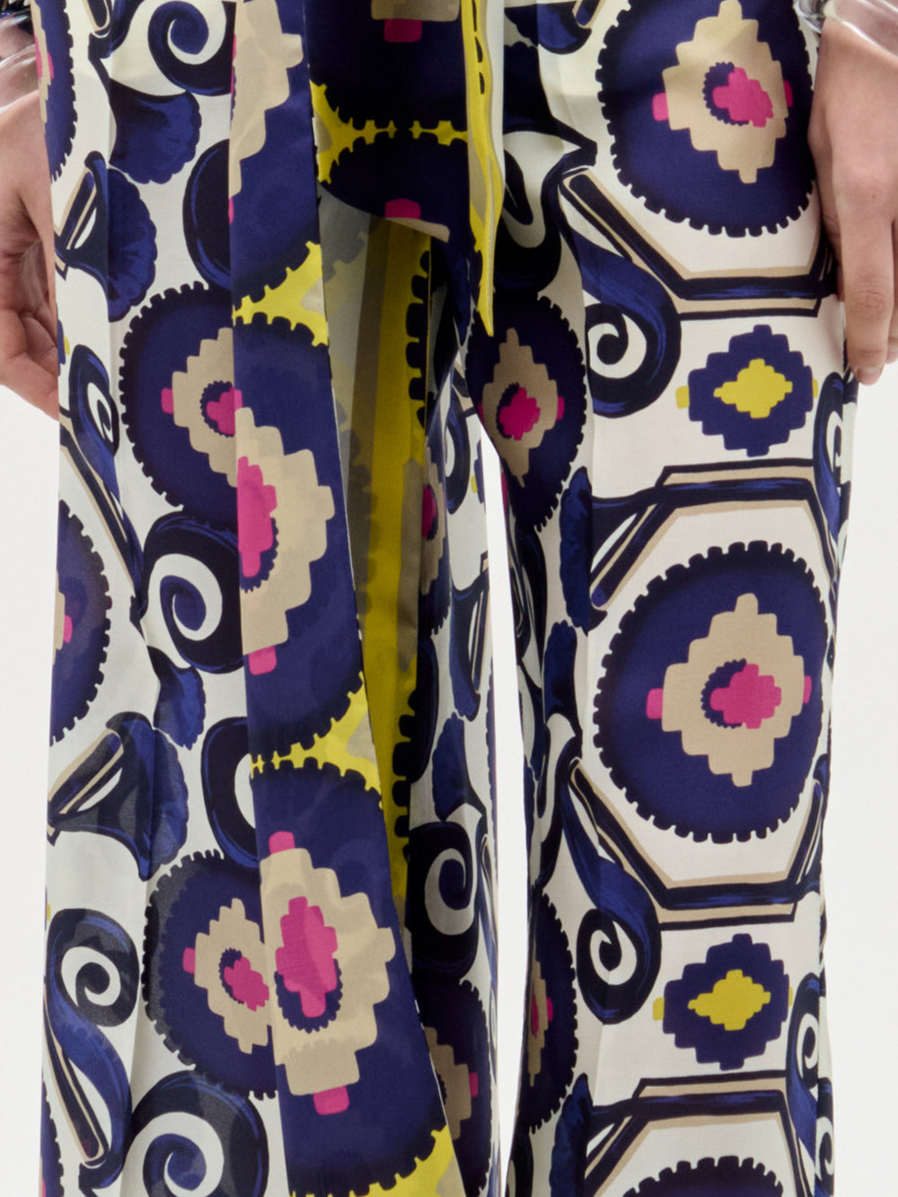 A woman poses against a plain background wearing the Emine Pant—wide-leg silk pants—layered under a long-sleeved, floor-length dress featuring a geometric multicolored pattern in blue, yellow, pink, and white for a chic modern look.