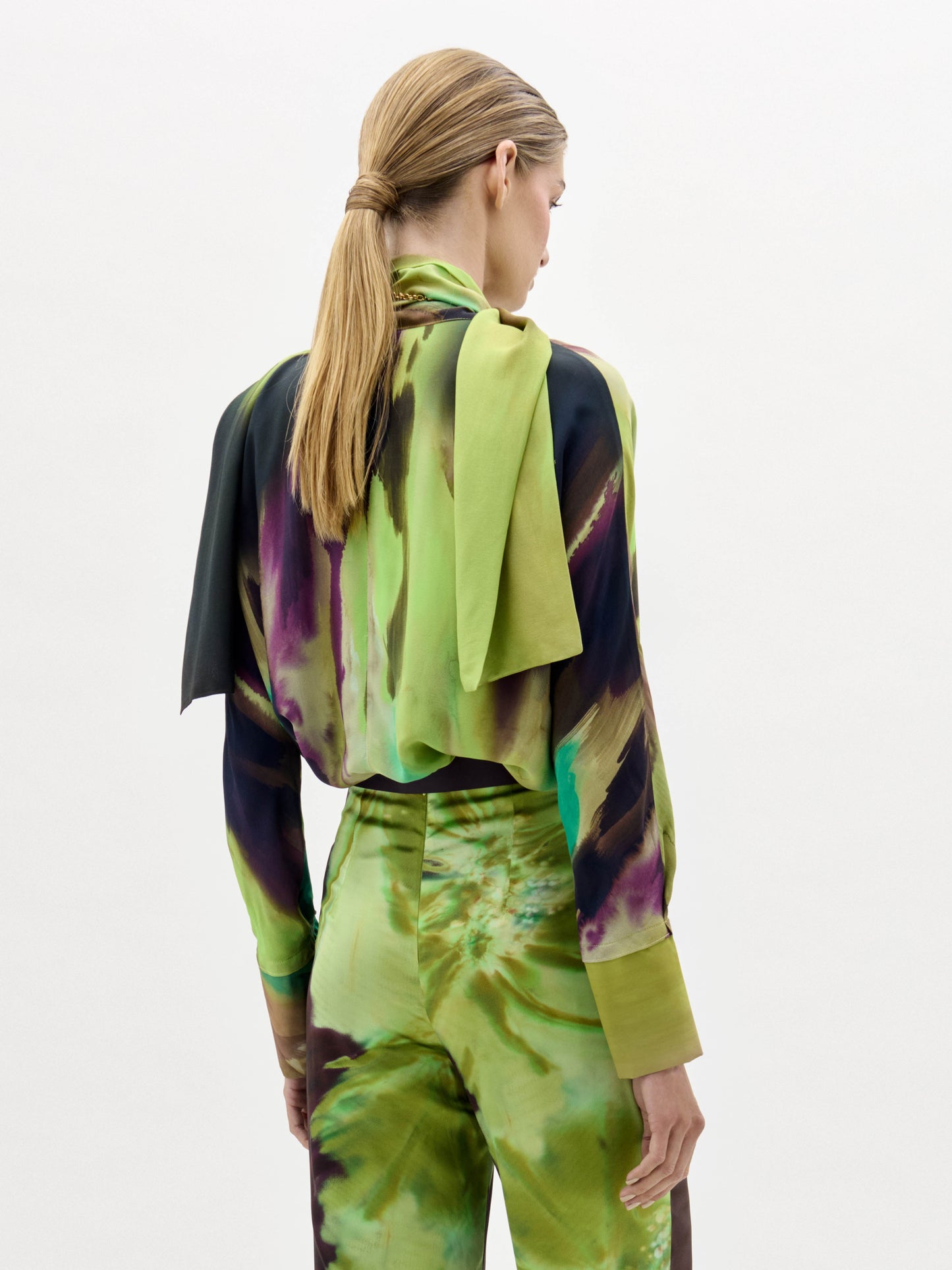 A woman stands against a plain white background wearing the Jaboca Blouse—a green, yellow, and brown tie-dye top styled with a wide belt and a scarf draped around her neck.