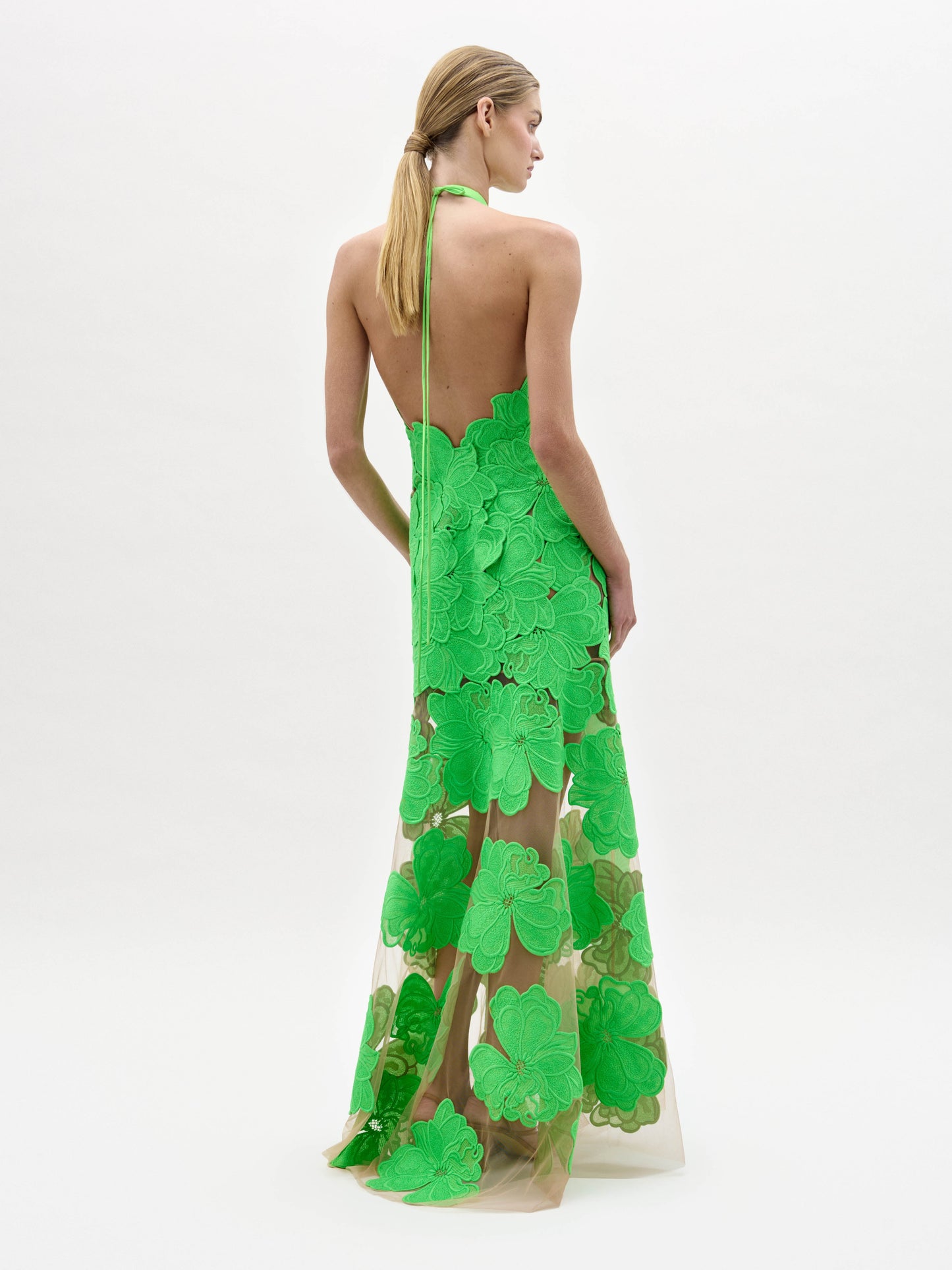 A woman models the Mab Dress, a sleeveless, floor-length gown featuring large green floral appliqués on a sheer nude base, set against a plain white background.