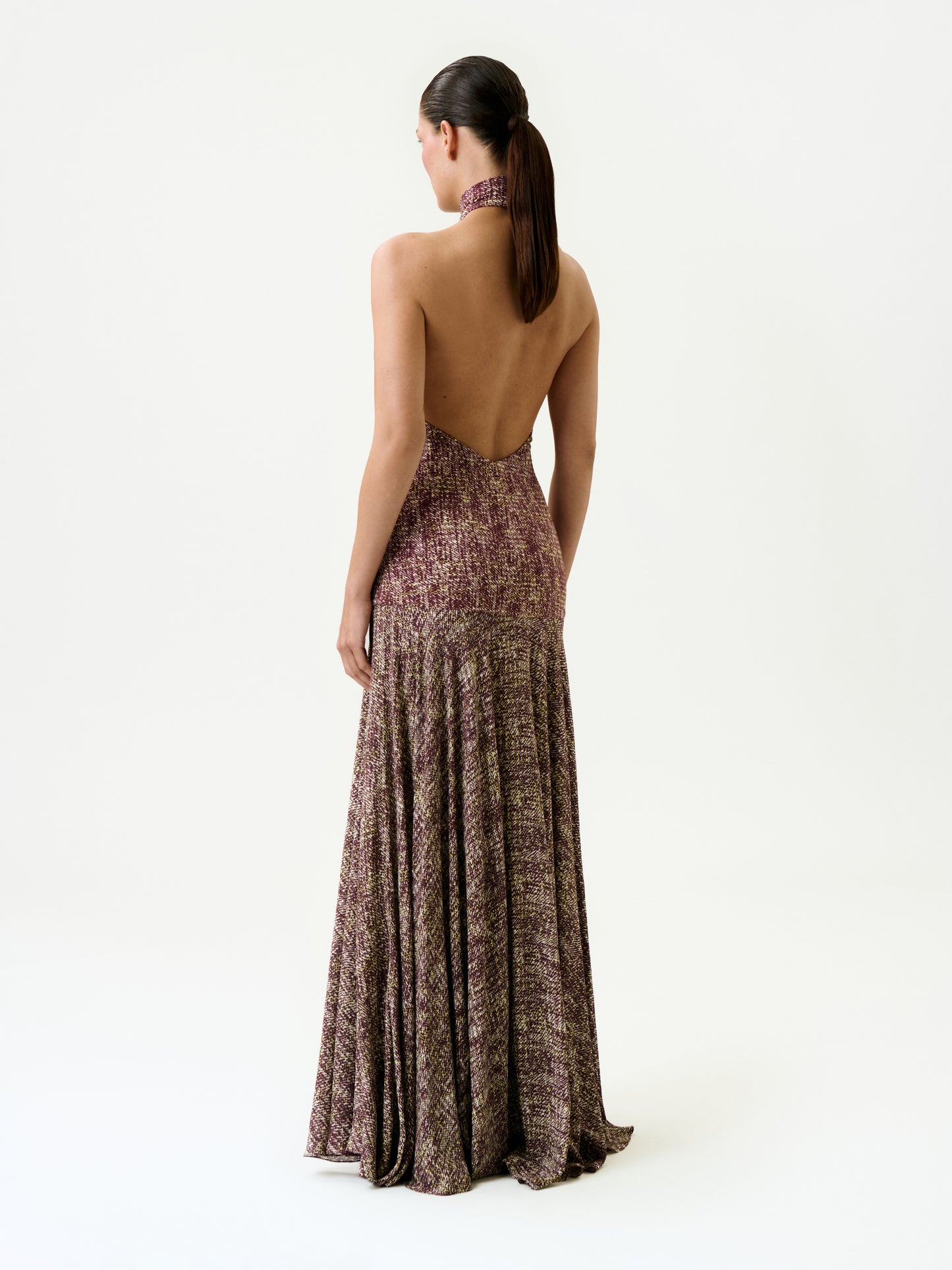 A woman with dark hair in a ponytail faces away from the camera, wearing the Leonore Dress—a halter-neck, backless maxi dress featuring a multicolored textured pattern, made in Colombia—against a plain white background.