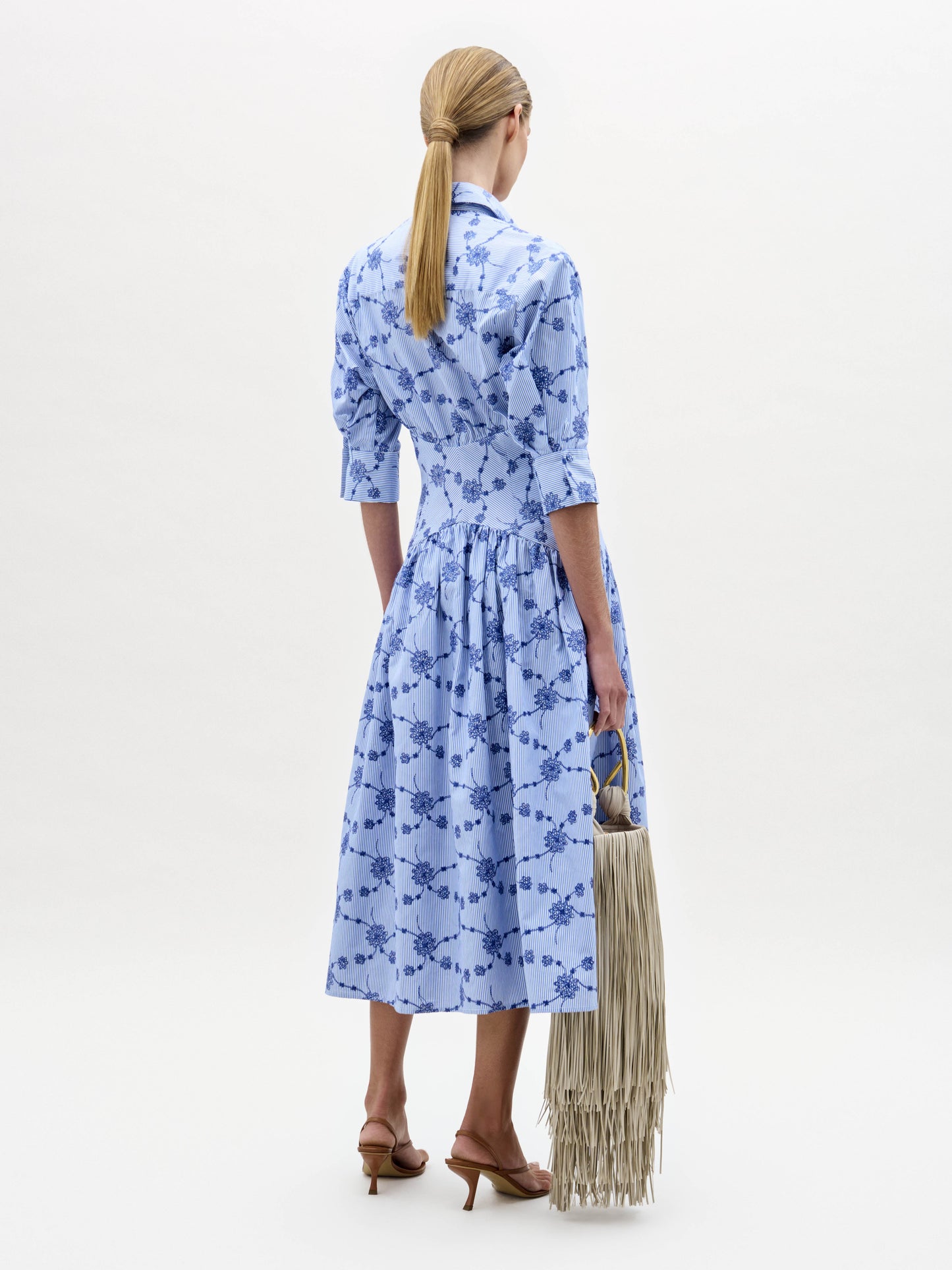 A woman stands against a plain white background wearing the Indy Dress—a light blue, patterned, collared midi dress with three-quarter sleeves—paired with brown sandals and a fringed handbag.