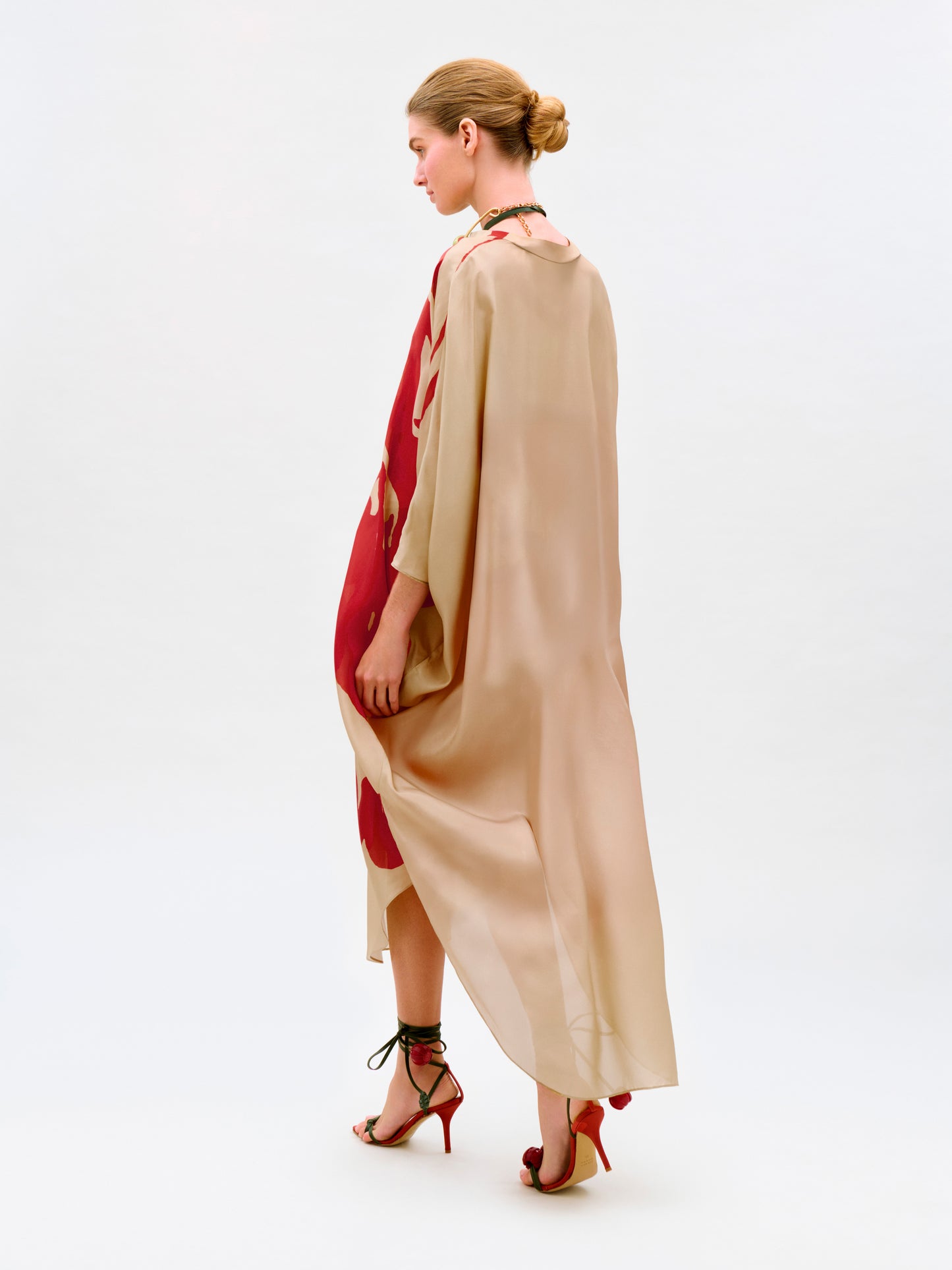 A woman with her hair in a bun wears the Elea Tunic, a beige and red floral maxi dress made from 100% Italian silk, paired with strappy red heels as she walks against a plain white background.