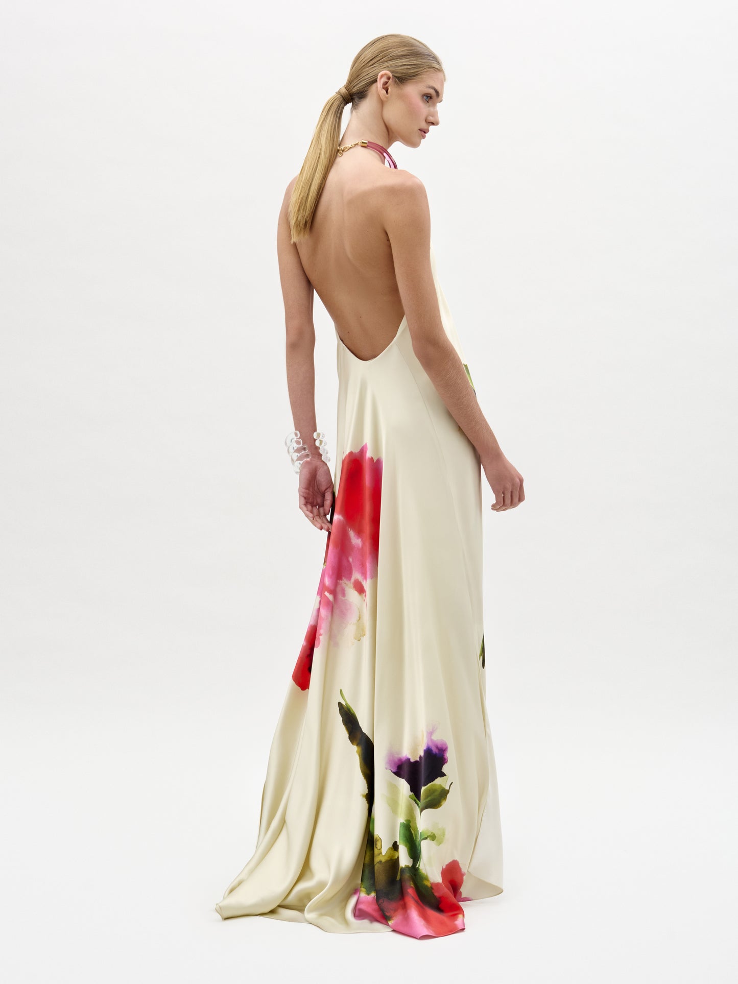 A woman with long, straight blonde hair in a low ponytail wears the cream-colored Jesse Dress—a backless maxi with a halter neckline and flowing train adorned with bold, watercolor floral prints—standing against a white background.