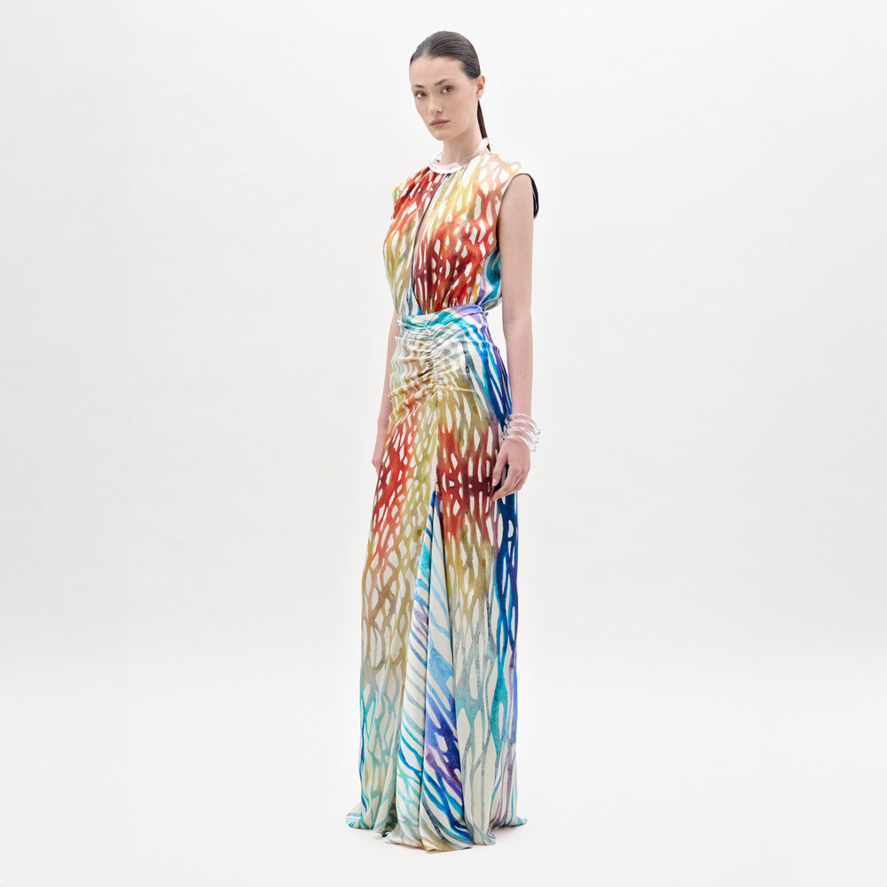 A woman stands against a plain background wearing the Samaira Dress, a sleeveless maxi with a ruched waist and a vibrant red, blue, yellow, and white abstract pattern.