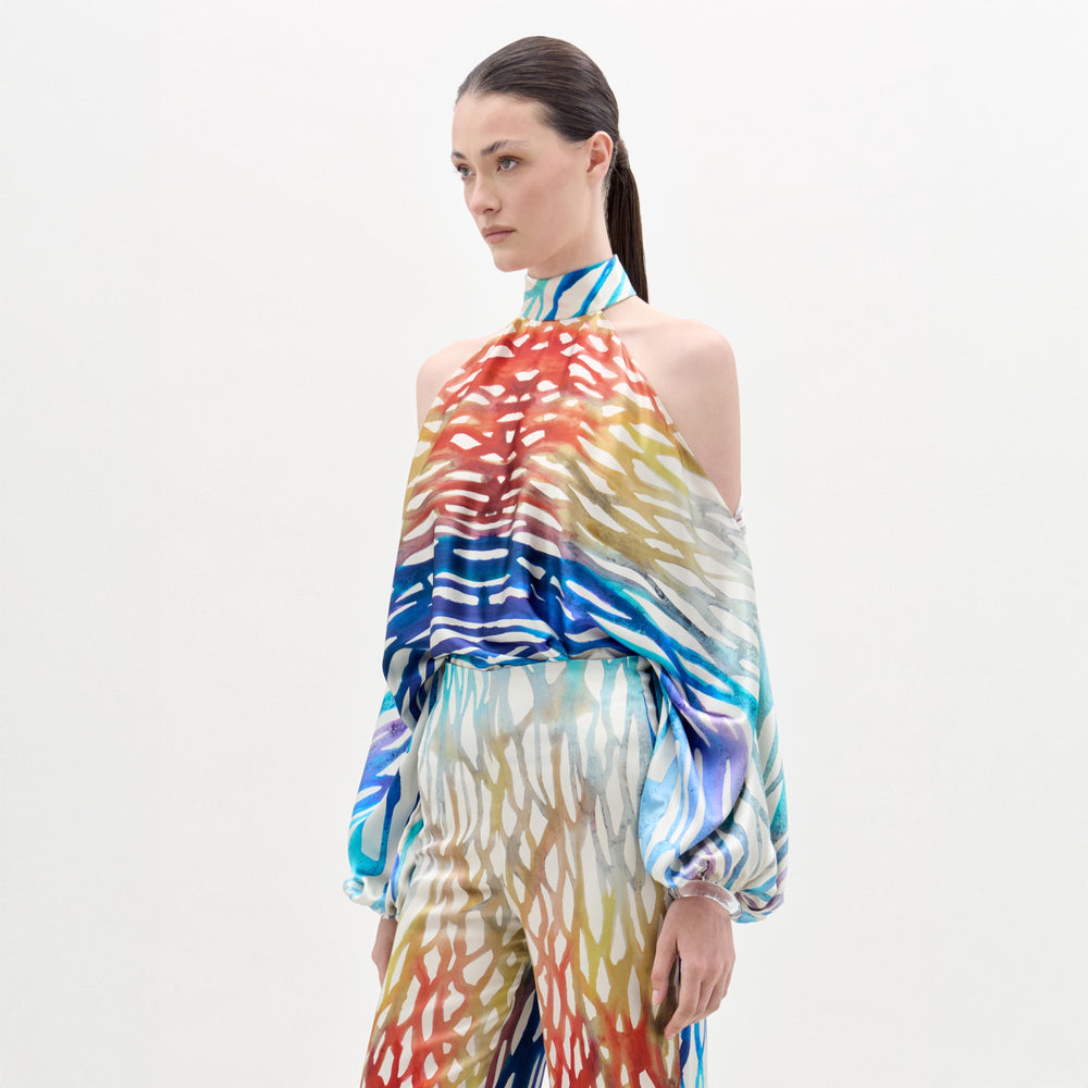 A woman models the Safira Blouse—an off-shoulder top featuring a colorful, abstract print in luxe Italian fabric—paired with matching wide-leg pants, set against a plain white background.