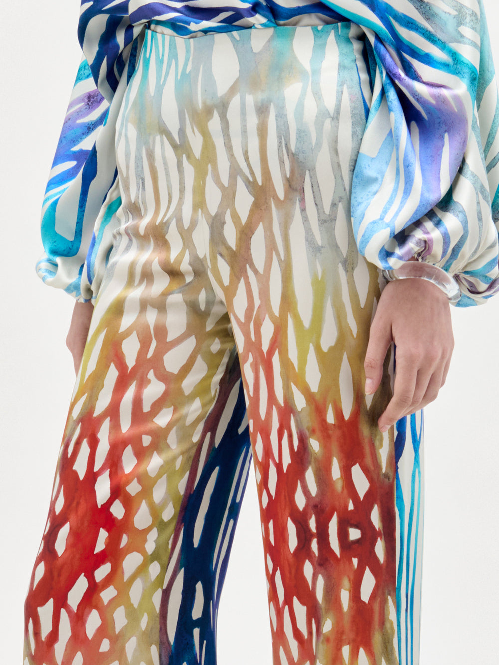 Woman poses against a plain background wearing the Andie Pant, a high-waisted, wide-leg style with colorful patterns, matching halter top, open shoulders, and long sleeves—all crafted from luxe Italian fabric.