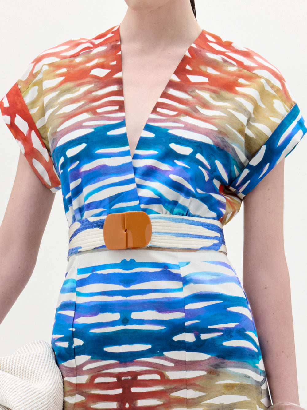 A woman stands against a white background wearing the Sakura Dress, a colorful patterned midi made from Italian fabric, paired with black heeled sandals and holding a large off-white bag.