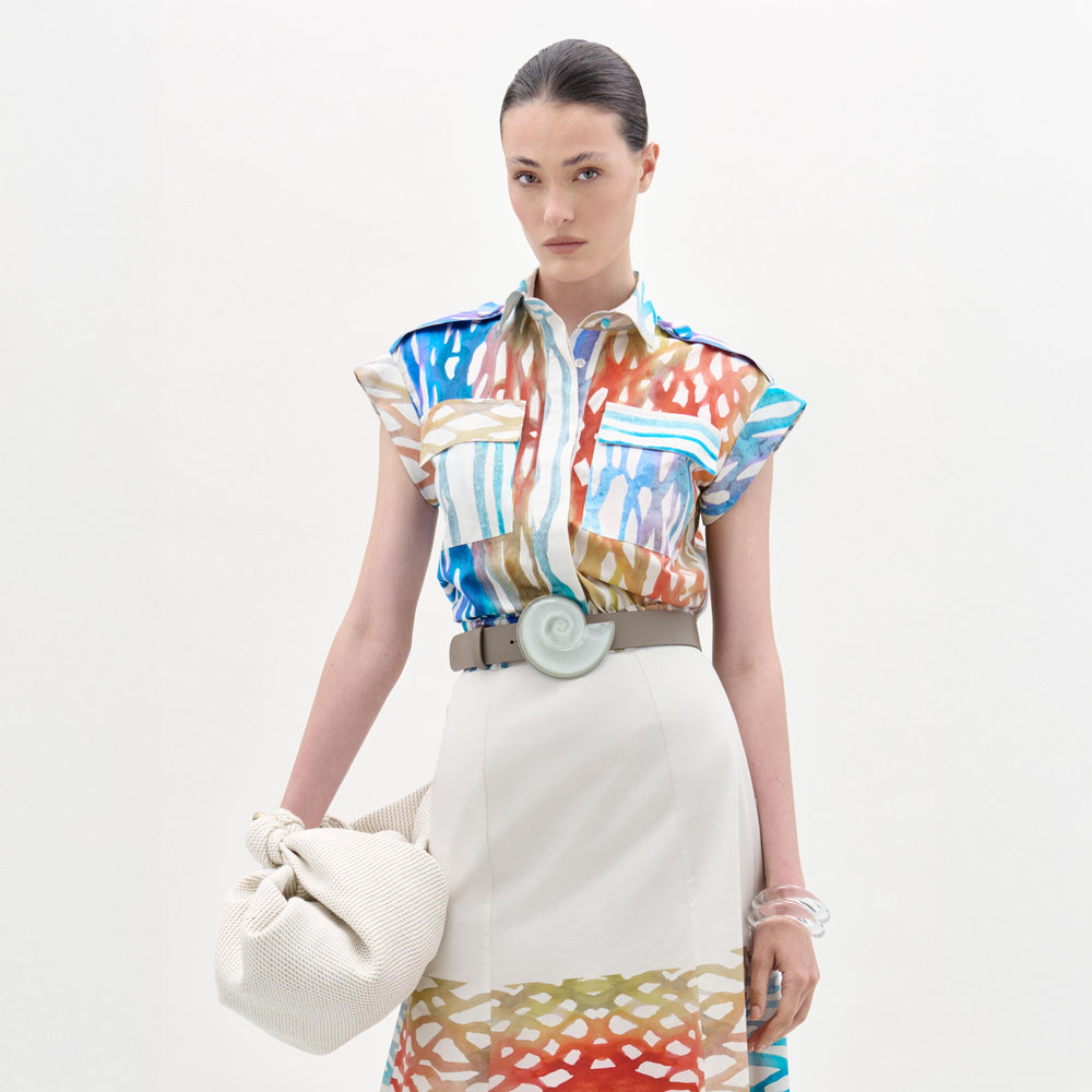 A woman stands against a white background wearing the Bara Blouse—crafted from 100% cotton Italian fabric—paired with a matching patterned beige skirt, wide spiral-buckle belt, and holding a textured cream bag.