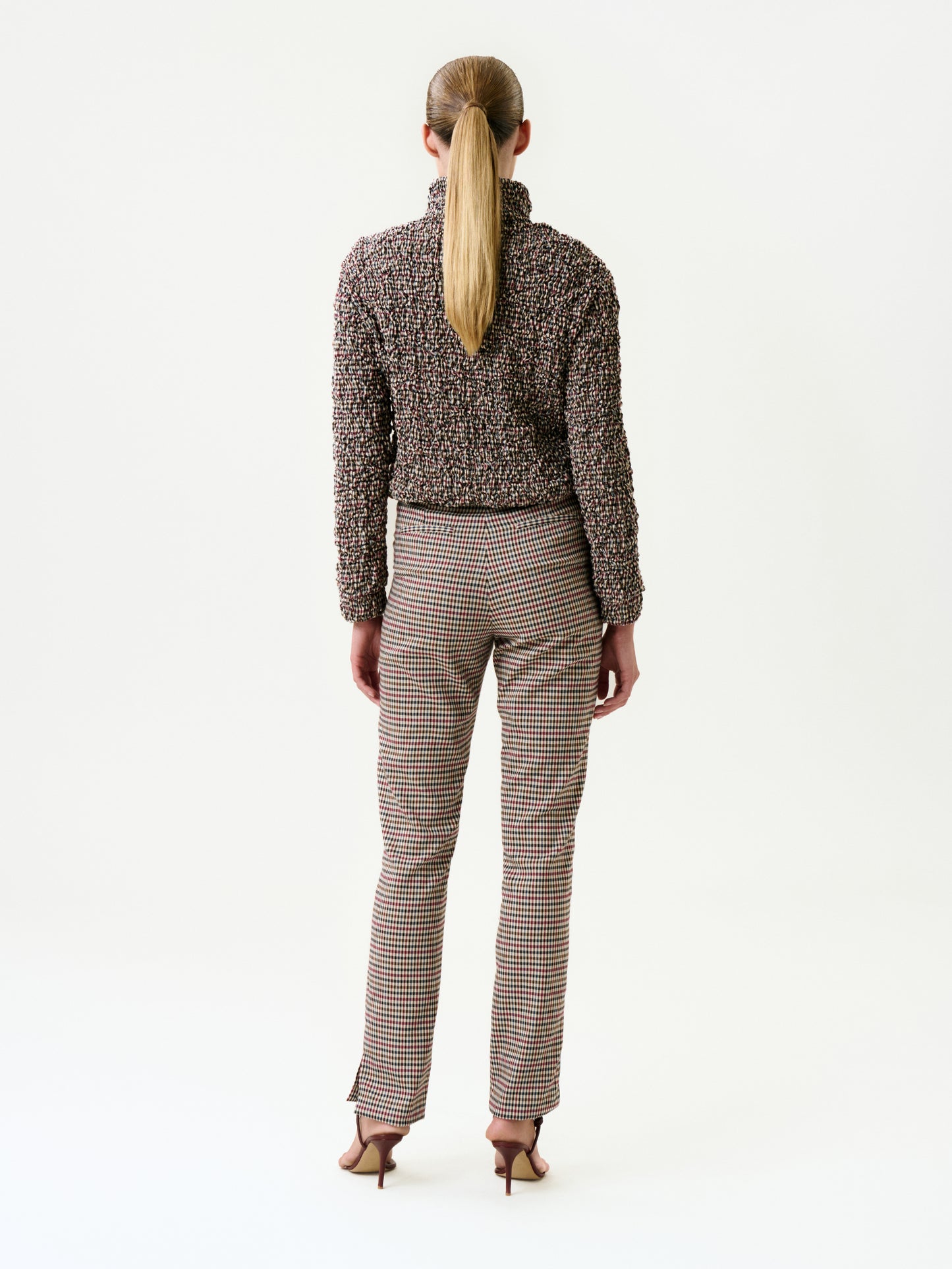A person with long straight hair in a low ponytail stands facing away, wearing the Luca Pant, a textured long-sleeve Italian-fabric top, and heeled sandals against a plain white background.