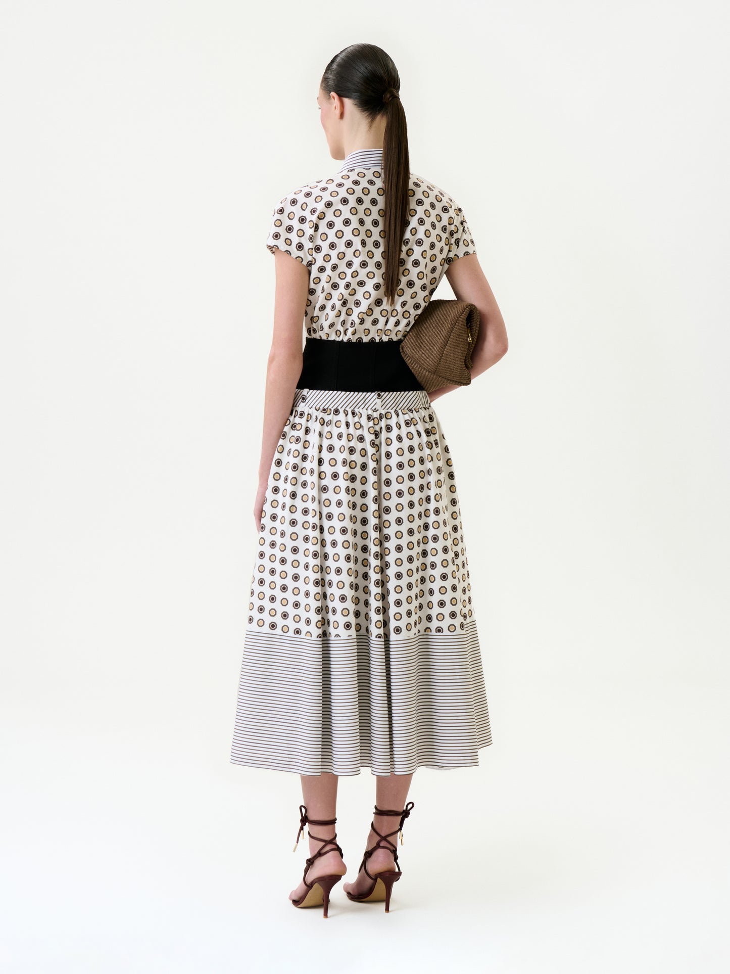 A woman with a low ponytail and strappy heels stands facing away on a plain white background, wearing the Odalis Dress—a patterned 100% cotton midi with a black waistband—and holding a textured brown clutch.