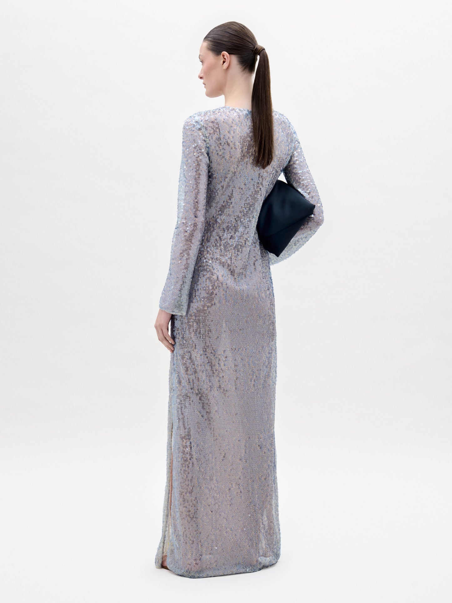 A woman stands against a plain background wearing the Nico Dress, a long-sleeved, floor-length silver sequin gown, and holding a black clutch.