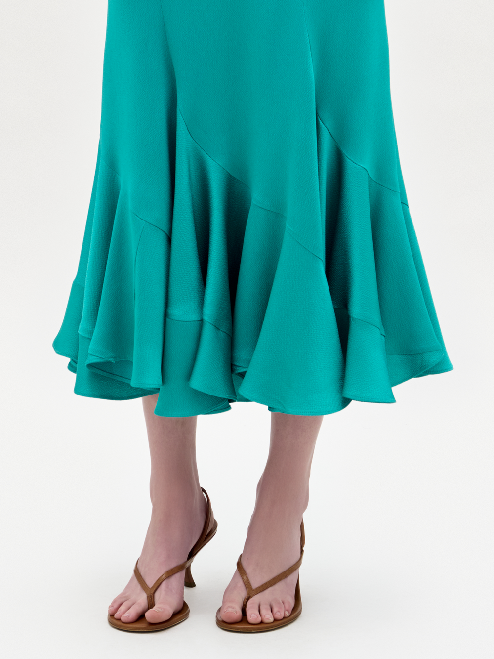 A woman stands against a plain background wearing the Marlene Skirt, styled with brown heeled sandals and holding a large pale pink clutch.