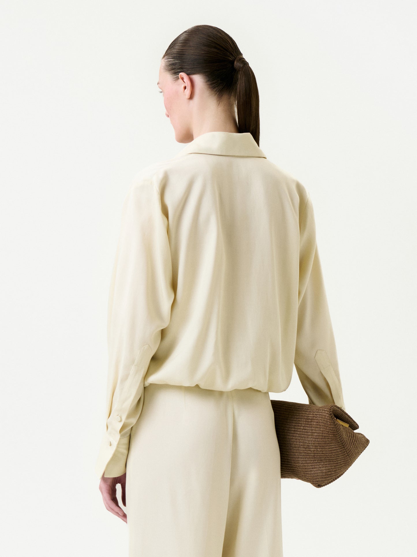 A woman with dark hair in a low ponytail is seen from behind, wearing the cream-colored Saula Blouse and pants, holding a textured brown clutch under her arm against a plain white background.