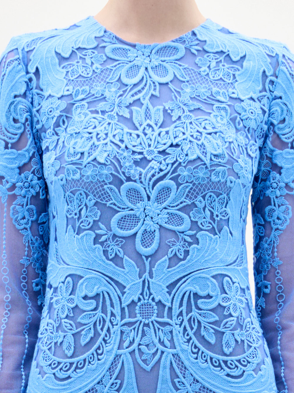 A woman stands facing forward in the Lacey Dress, a long, fitted floral lace gown with intricate patterns, long sleeves, and a beautifully tailored bodice, set against a plain light background.