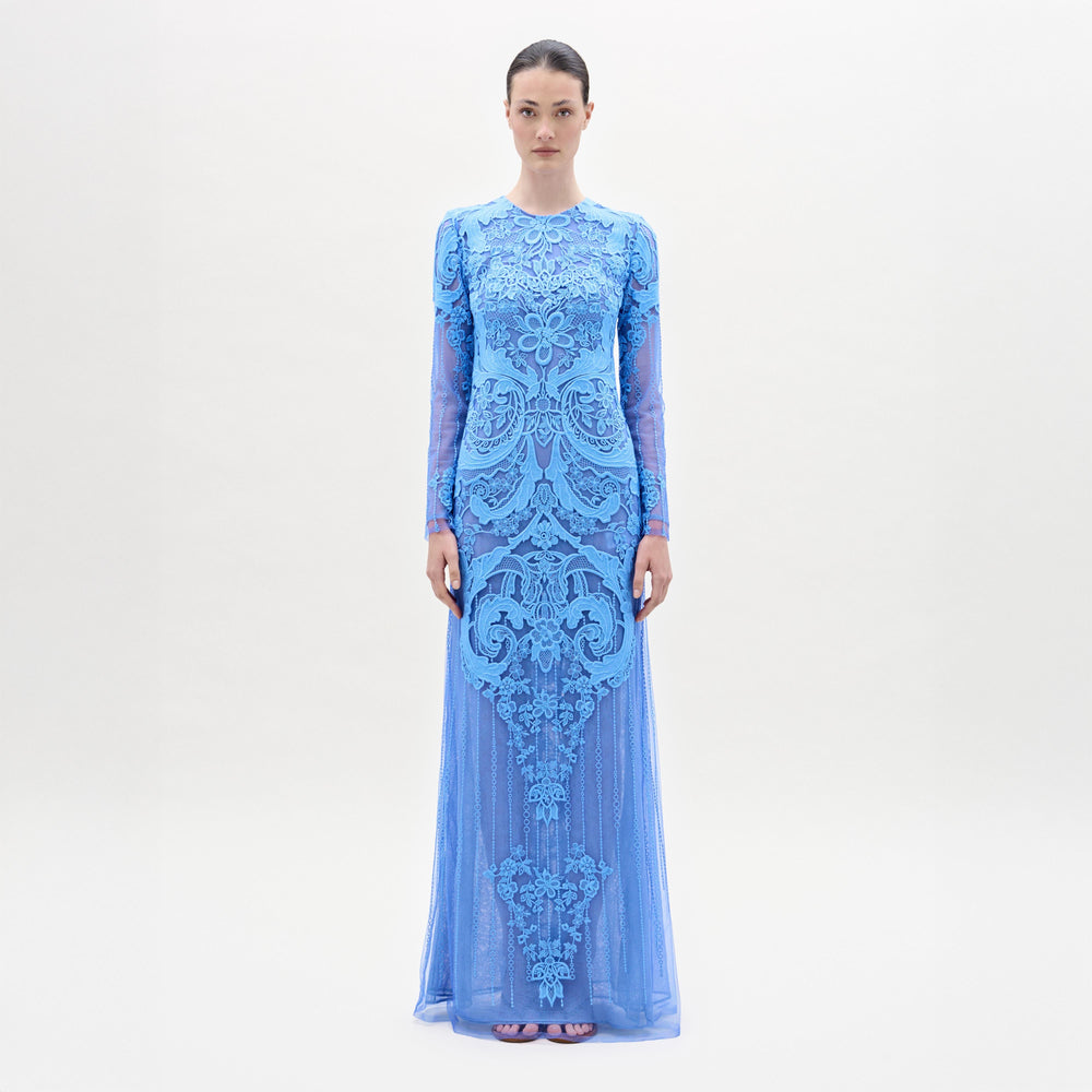 A woman stands facing forward in the Lacey Dress, a long, fitted floral lace gown with intricate patterns, long sleeves, and a beautifully tailored bodice, set against a plain light background.