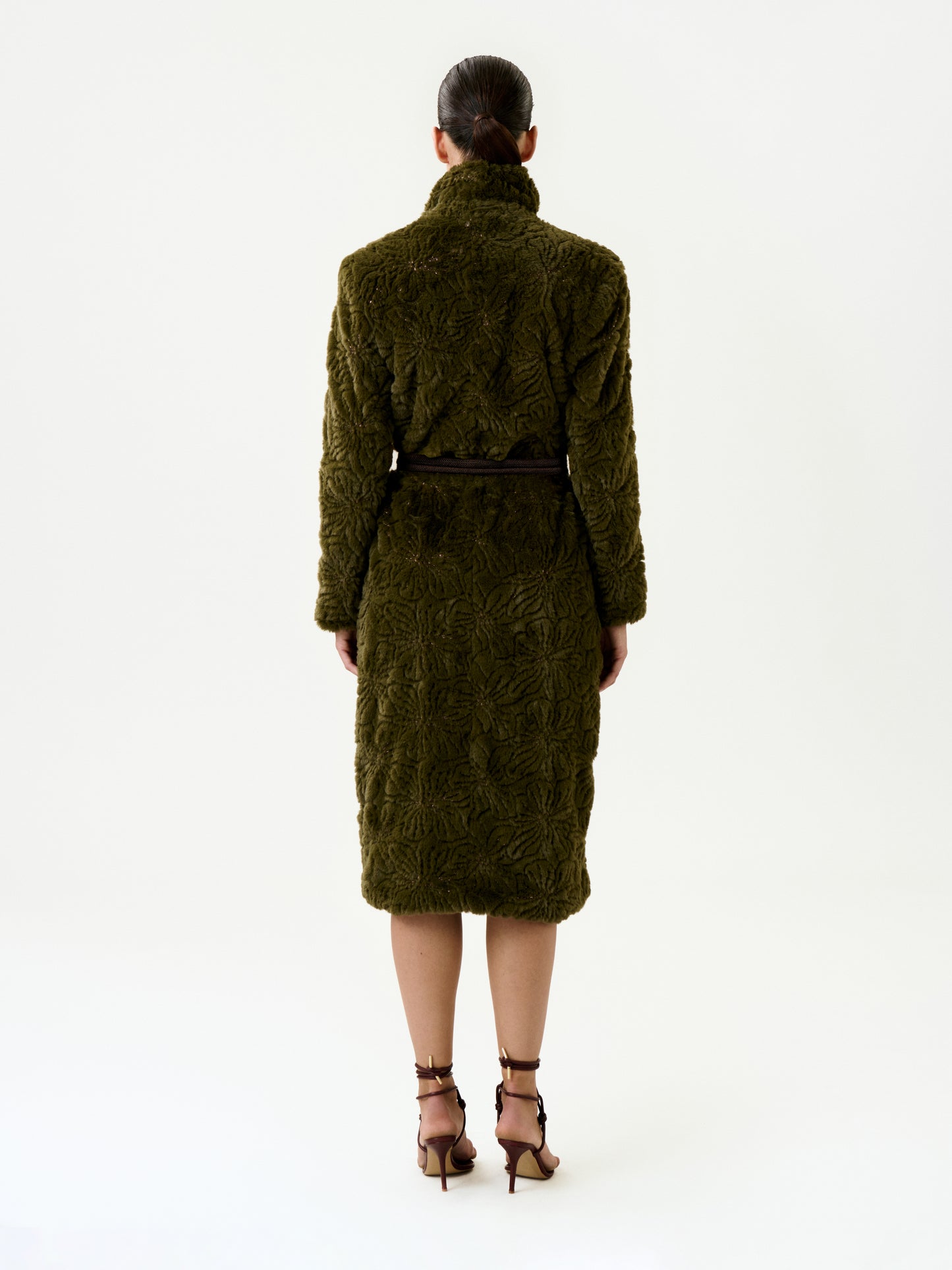 A woman stands facing away against a plain white background, wearing the Emerson Trench—a knee-length, textured olive green coat crafted from luxurious Italian fabric in an oversized fit—cinched at the waist with a black belt and paired with brown heels.