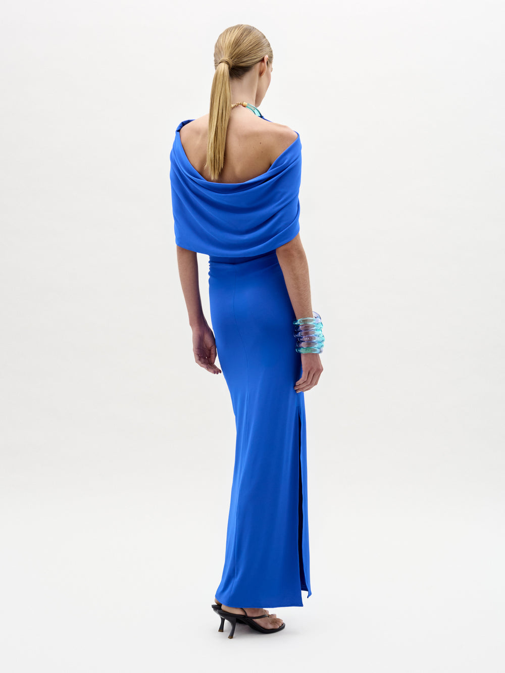Woman in the Odele Dress, a fitted, floor-length bright blue maxi with cape-like detail and halter neckline, stands against a white background wearing heeled sandals and matching blue bracelets.