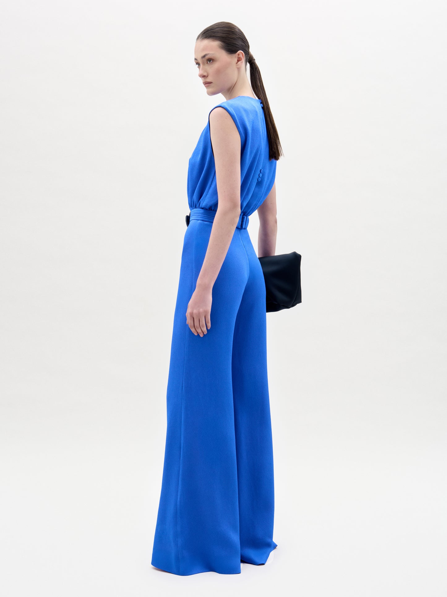 A woman with straight brown hair pulled back stands sideways against a white background, wearing the Noel Jumpsuit—a sleeveless, wide-leg style made from fine Italian textile—and holds a black clutch.