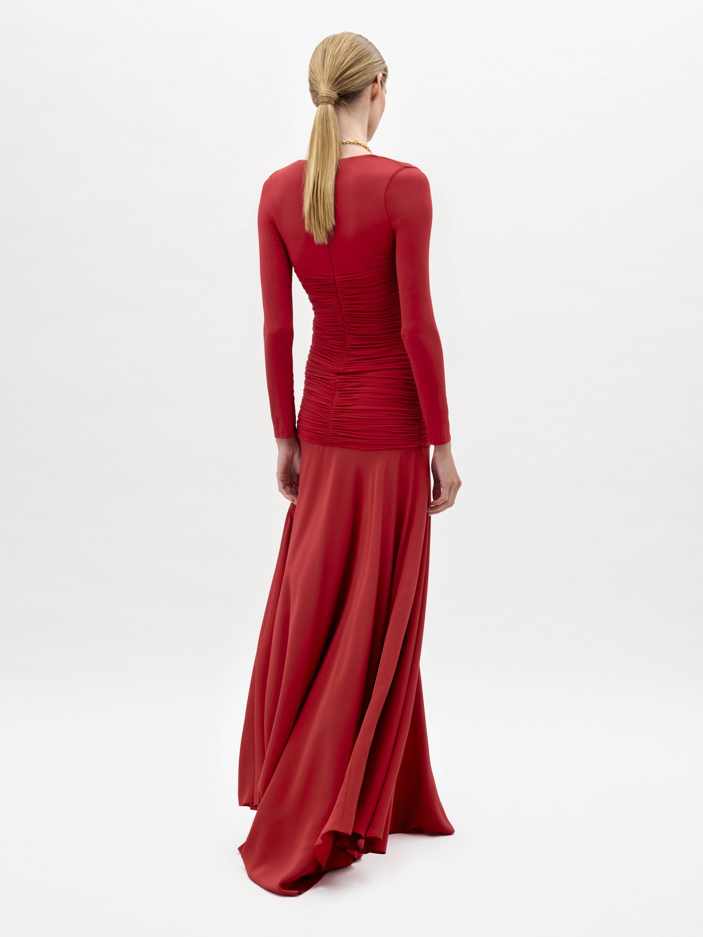 A woman models the Lina Dress, a long-sleeved, floor-length red gown with a deep V-neckline, paired with a gold necklace, against a plain white background.