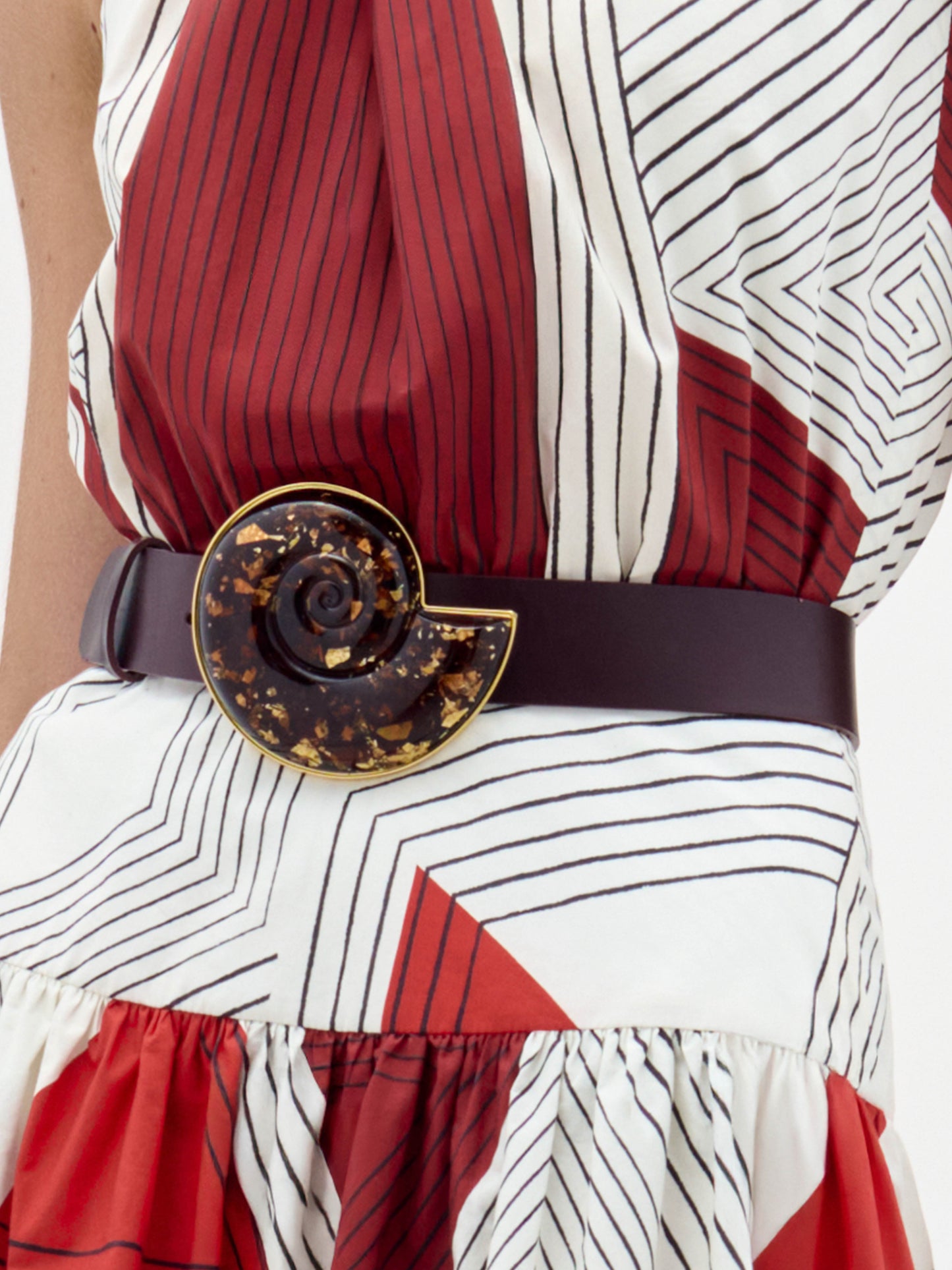 The Nautilus Belt is a dark maroon accessory with a large, glossy, spiral-shaped buckle adorned with amber and gold flecks.