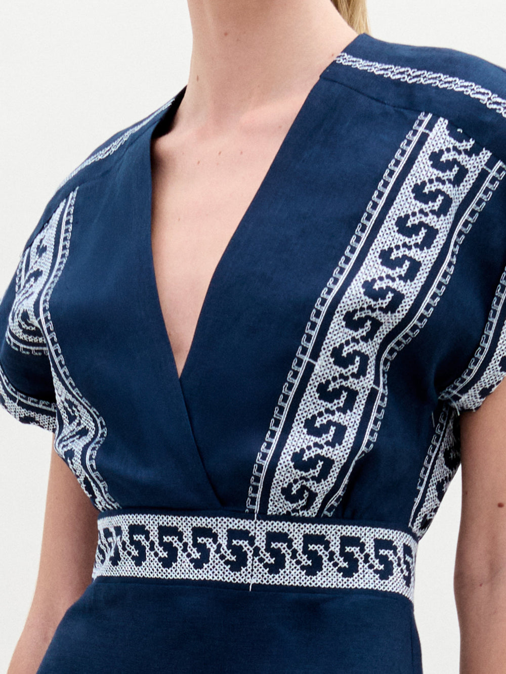 A woman stands against a plain background wearing the Ady Dress, a navy blue embroidered linen midi with white patterns, gold sandals, and holding a light-colored handbag.