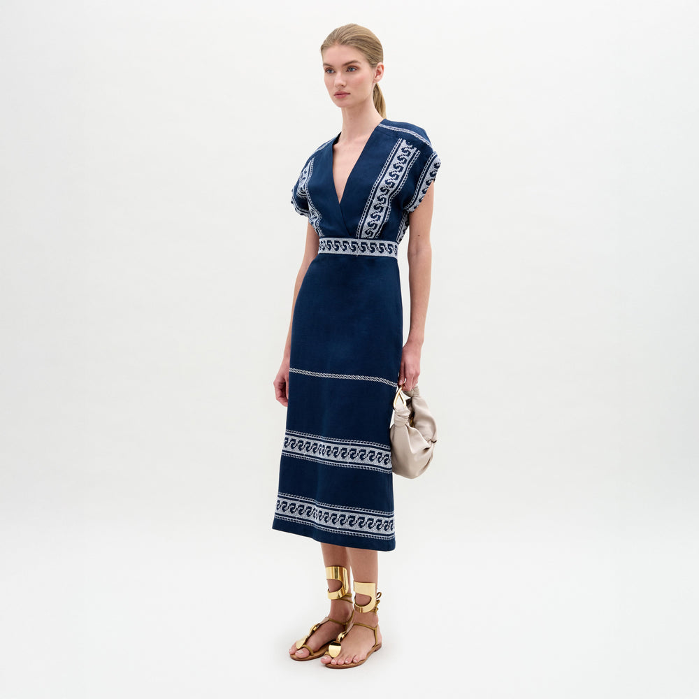A woman stands against a plain background wearing the Ady Dress, a navy blue embroidered linen midi with white patterns, gold sandals, and holding a light-colored handbag.