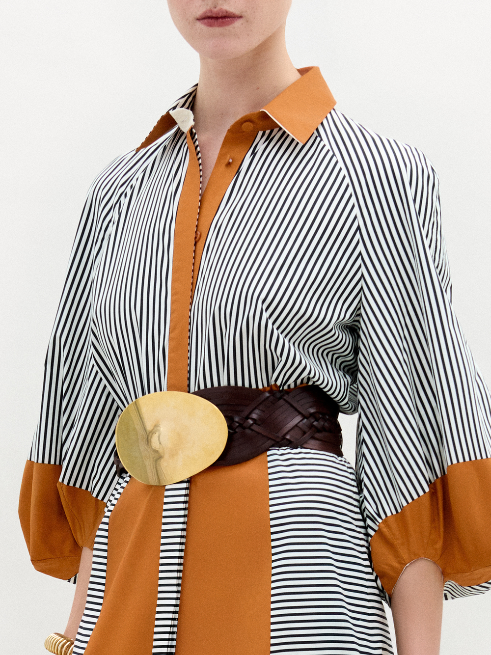 A woman stands against a white background wearing the Agostina Dress—striped and color-blocked, crafted from Italian fabric—paired with high brown boots, a wide belt, and holding a large dark bag.