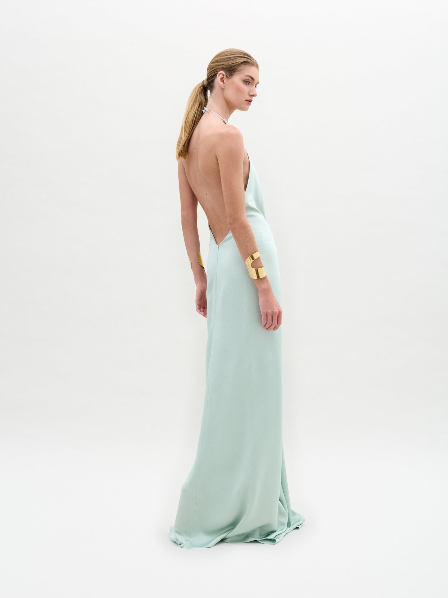 A woman wears the Aiko Dress, a light blue, floor-length maxi made from Italian fabric with a deep V-neck and central clasp, styled with gold bracelets against a plain white background.