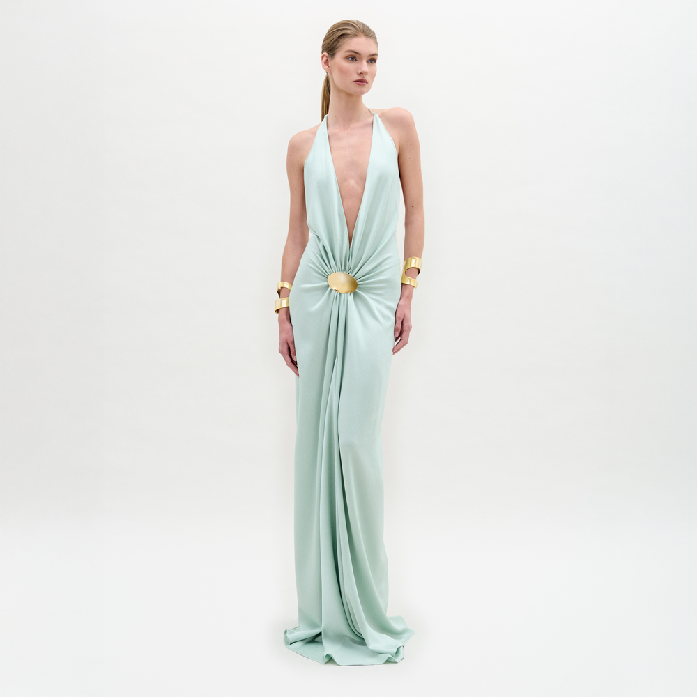 A woman wears the Aiko Dress, a light blue, floor-length maxi made from Italian fabric with a deep V-neck and central clasp, styled with gold bracelets against a plain white background.