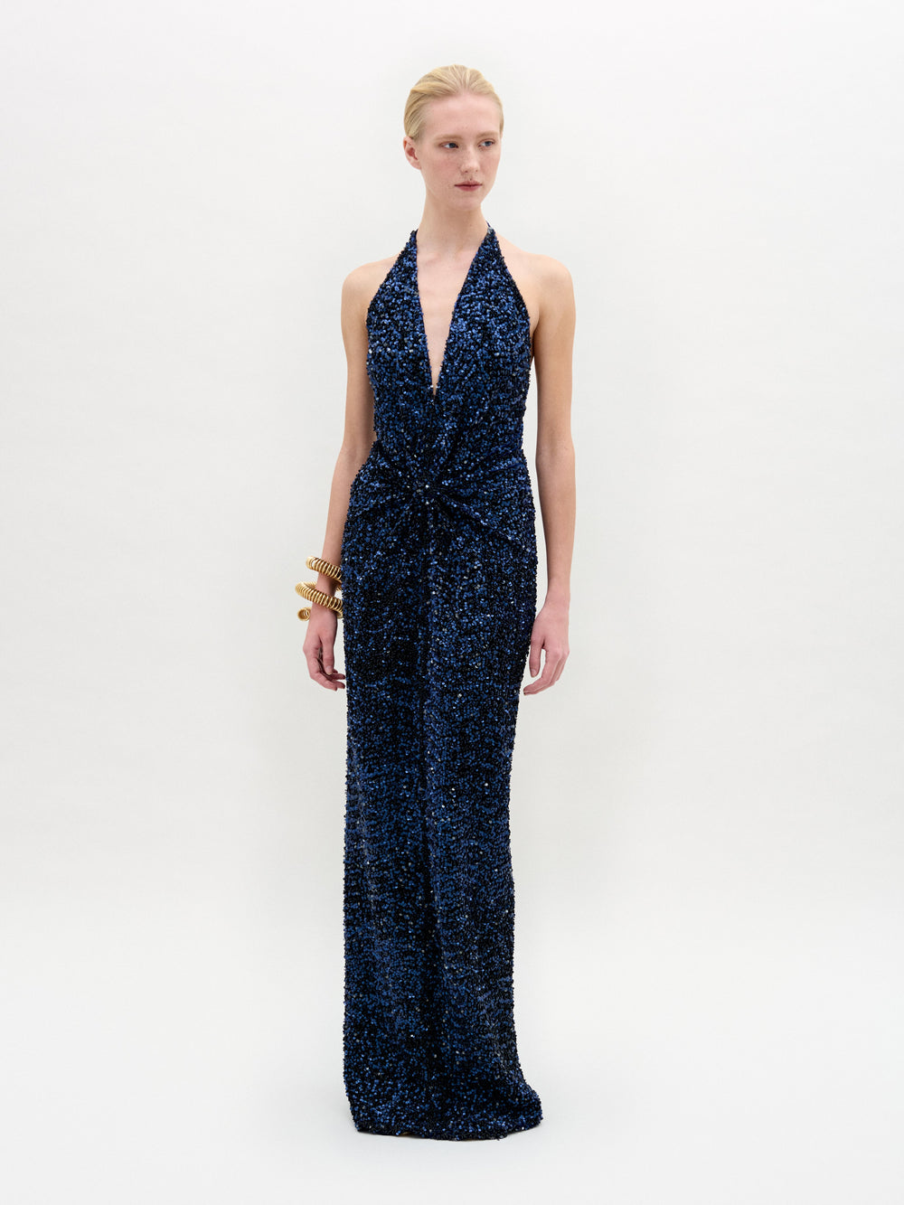 A person dressed in a sleeveless Aiko Dress—a deep blue Atelier creation adorned with sequins and featuring an elegant halter neckline—is accessorized with gold bangles while standing against a plain background.