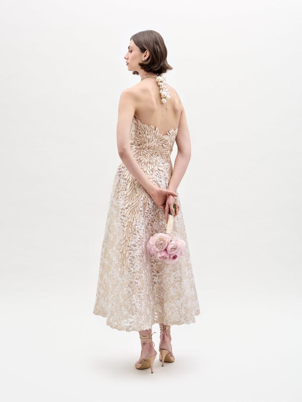 A woman in the strapless Alejandra Dress holds pink flowers. She wears pearl jewelry and beige heels, standing against a plain white background.