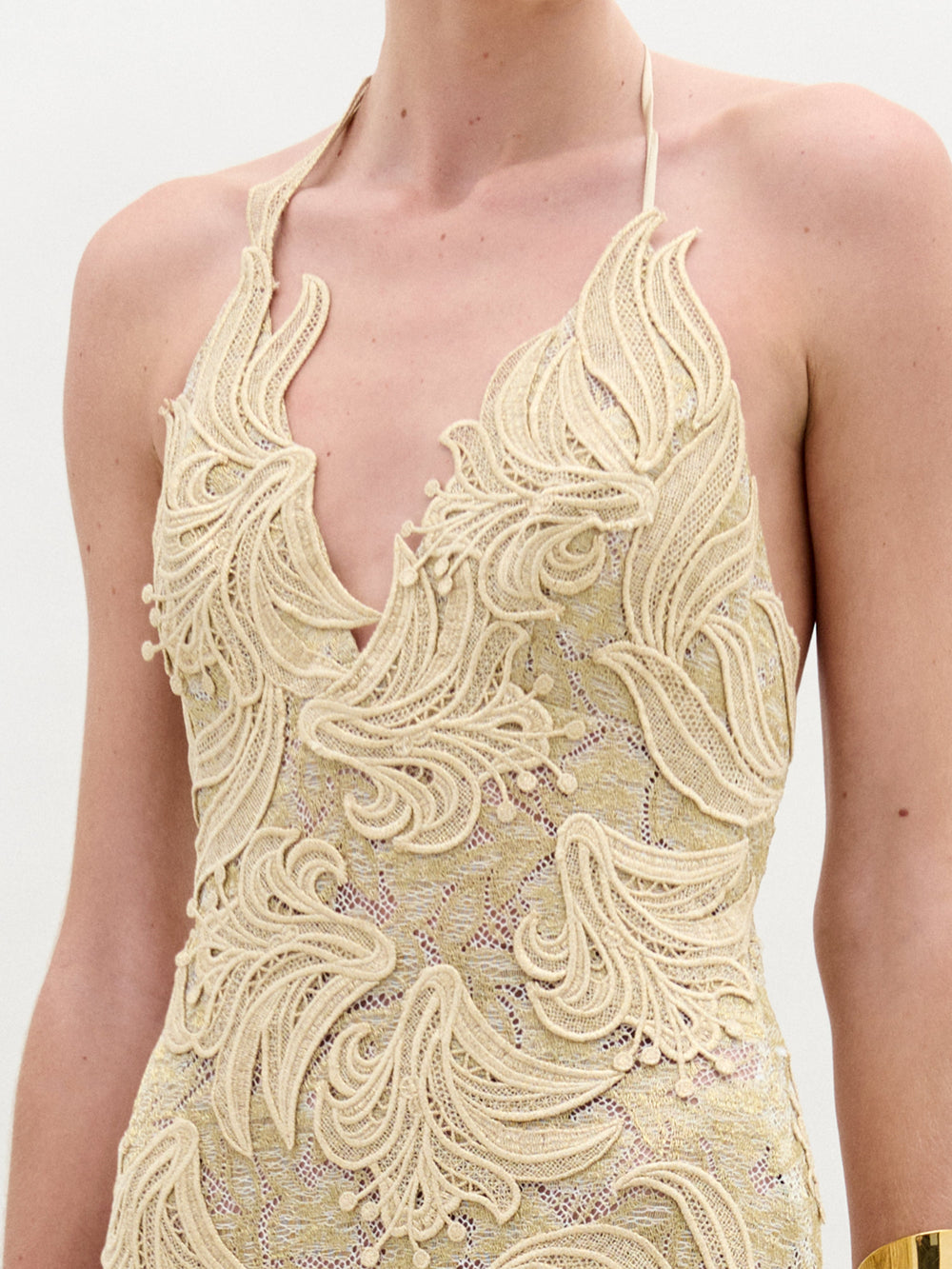 A woman wears the Aurora Dress—floor-length, sleeveless, crafted from gold lace with a fitted silhouette and wavy patterns—paired with gold cuff bracelets, standing against a plain white background.