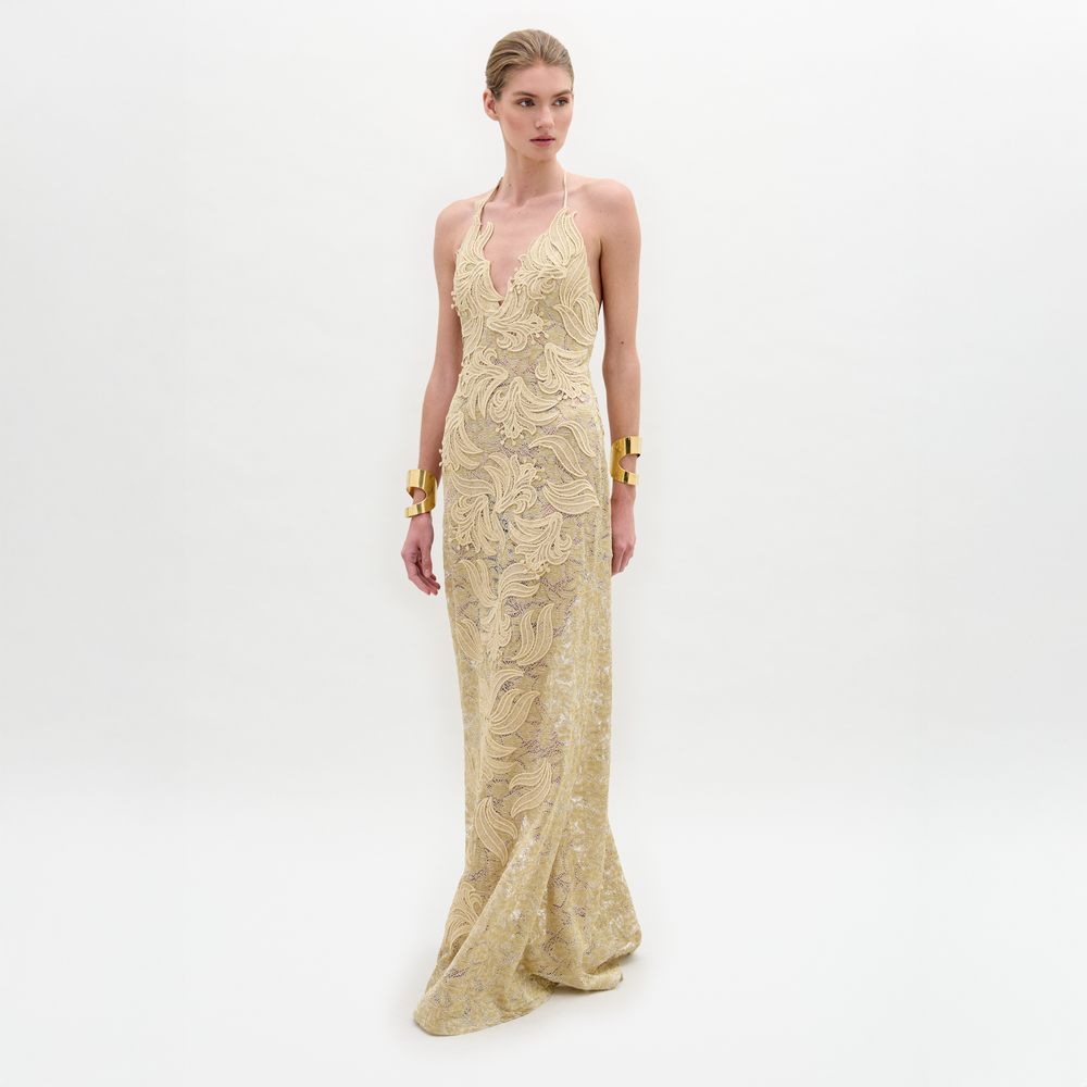 A woman wears the Aurora Dress—floor-length, sleeveless, crafted from gold lace with a fitted silhouette and wavy patterns—paired with gold cuff bracelets, standing against a plain white background.