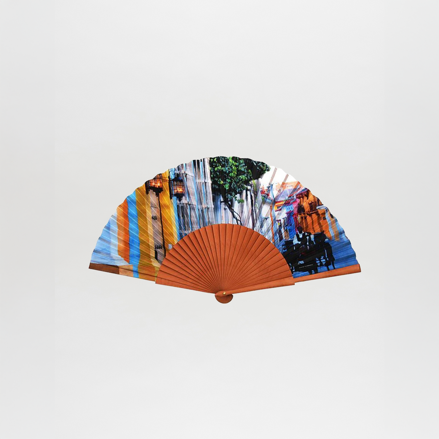 The Abanico Cochero is a handheld folding fan with a wooden frame and colorful fabric featuring a lively street scene with people, trees, and buildings, shown against a white background.