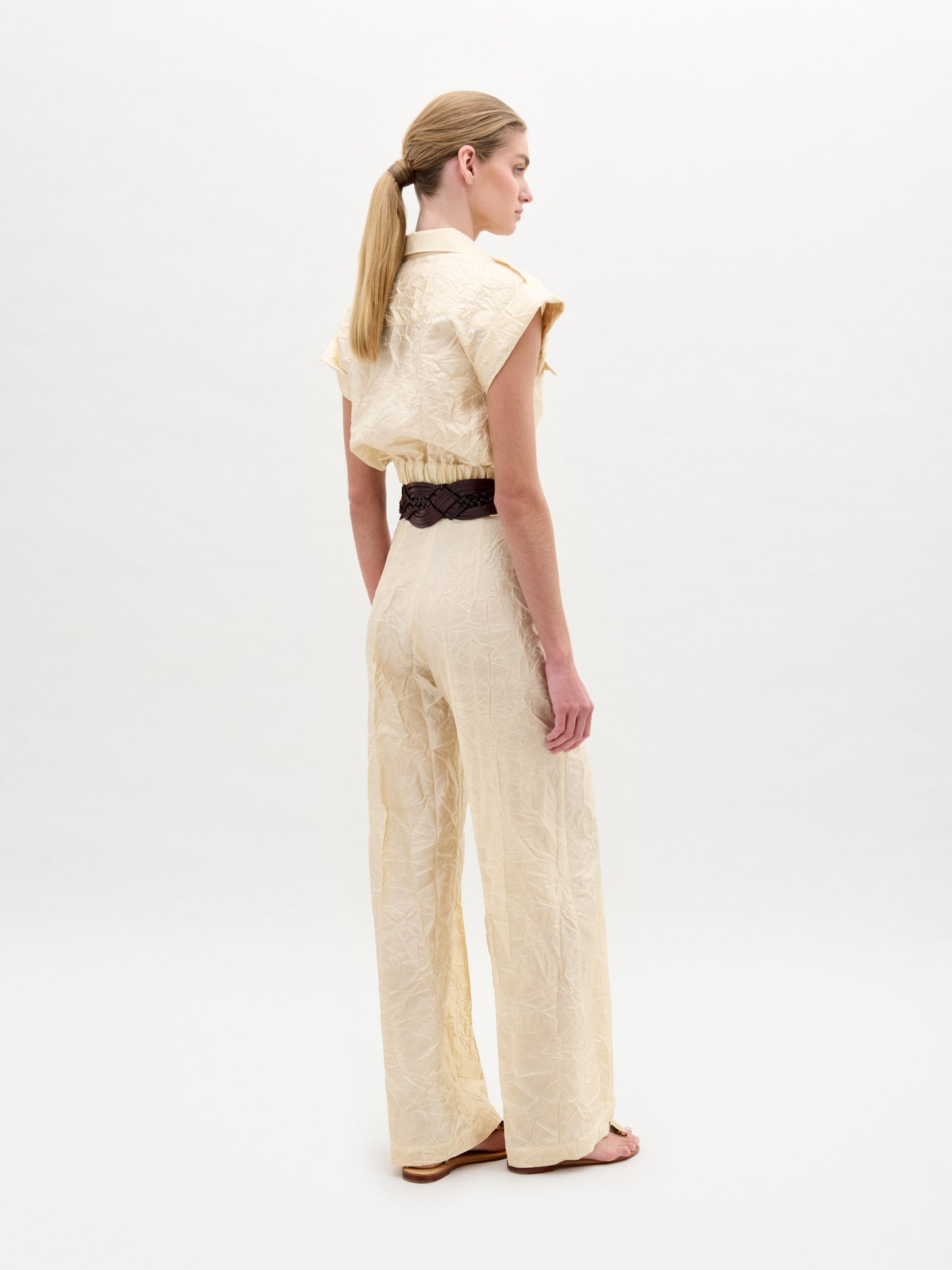 A person stands against a plain background wearing the Emine Pant, a beige textured wide-leg silhouette crafted from Italian fabric, styled with short sleeves, a wide black belt with gold buckle, and sandals.
