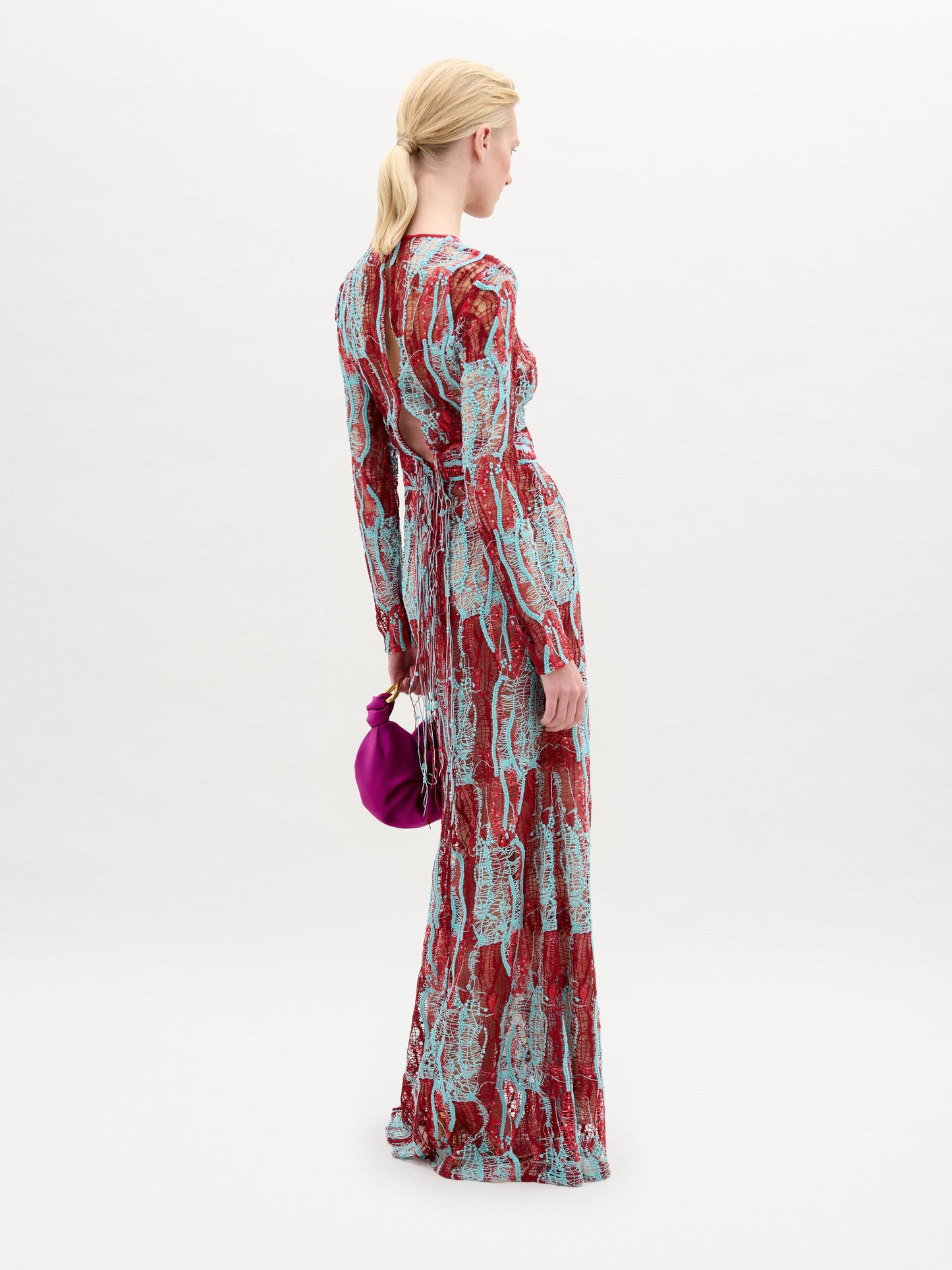 A person stands against a plain background wearing the Barisa Dress, a long, patterned red and blue fitted dress, and holding a small purple handbag.