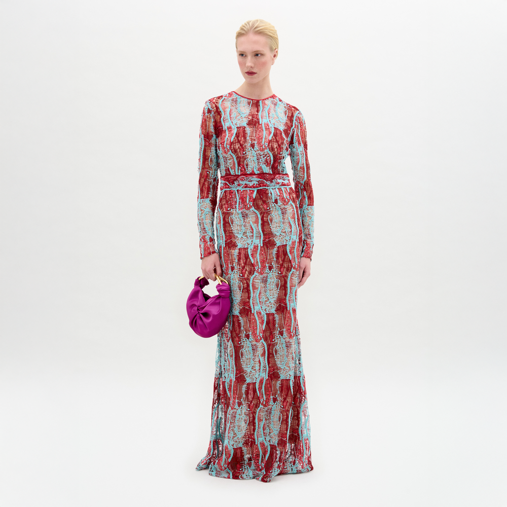 A person stands against a plain background wearing the Barisa Dress, a long, patterned red and blue fitted dress, and holding a small purple handbag.