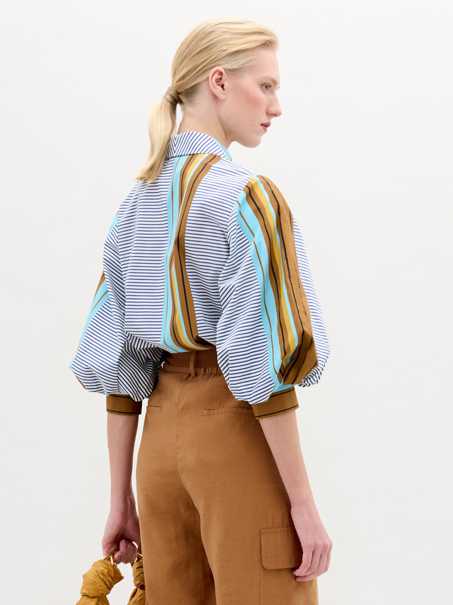 A person stands against a plain background wearing the oversized fit Belkis Blouse with balloon sleeves and stripes, paired with tan high-waisted pants, and holding a tan accessory in their left hand.